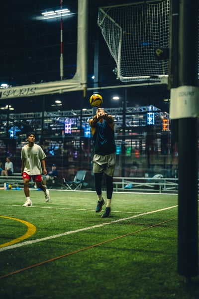 A player in a dark blue top and grey shorts jumps with arms extended toward the ball.