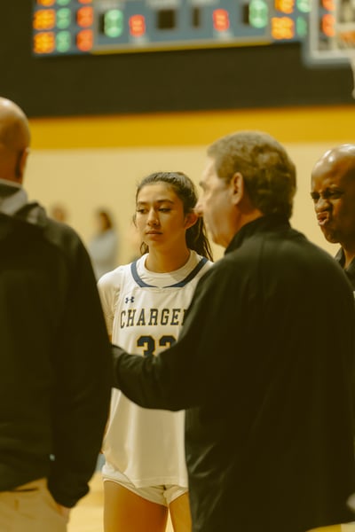 A player in white stands with coaches in black uniforms on a basketball court.