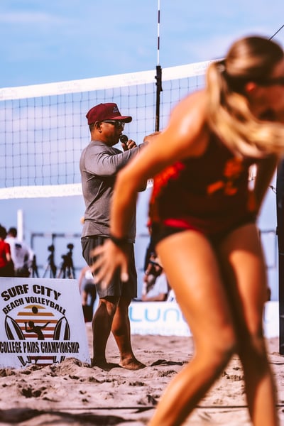 A person in a maroon hat speaks into a microphone on a beach court.