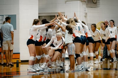 A team in white uniforms huddles with hands raised, one player in front crouches.