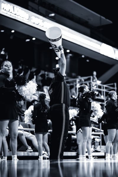 A player in black holds a megaphone overhead while players in black hold pom-poms.