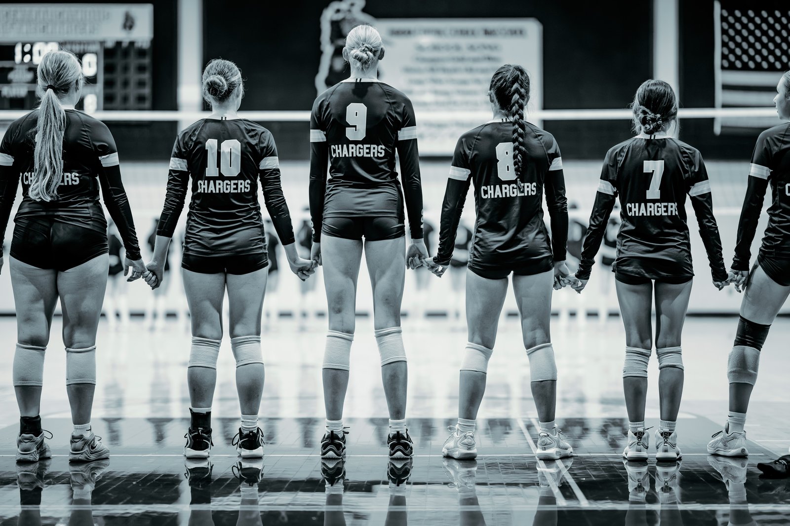 Players in black jerseys with white numbers hold hands on the court: number 10, number 9, number 8, and number 7.