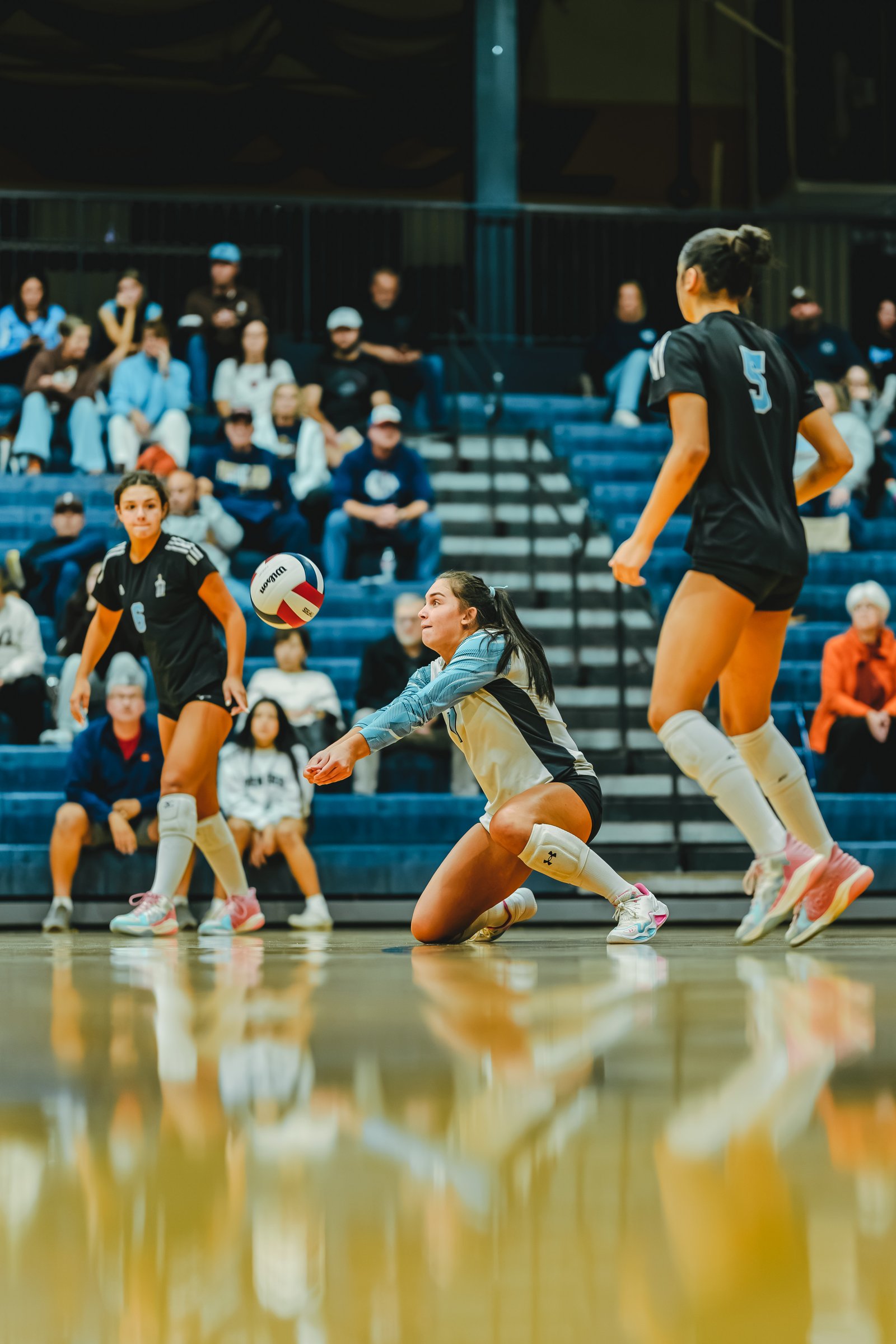 A player in a light blue jersey dives for a volleyball, while players in black jerseys with numbers 5 and 6 are on the court.