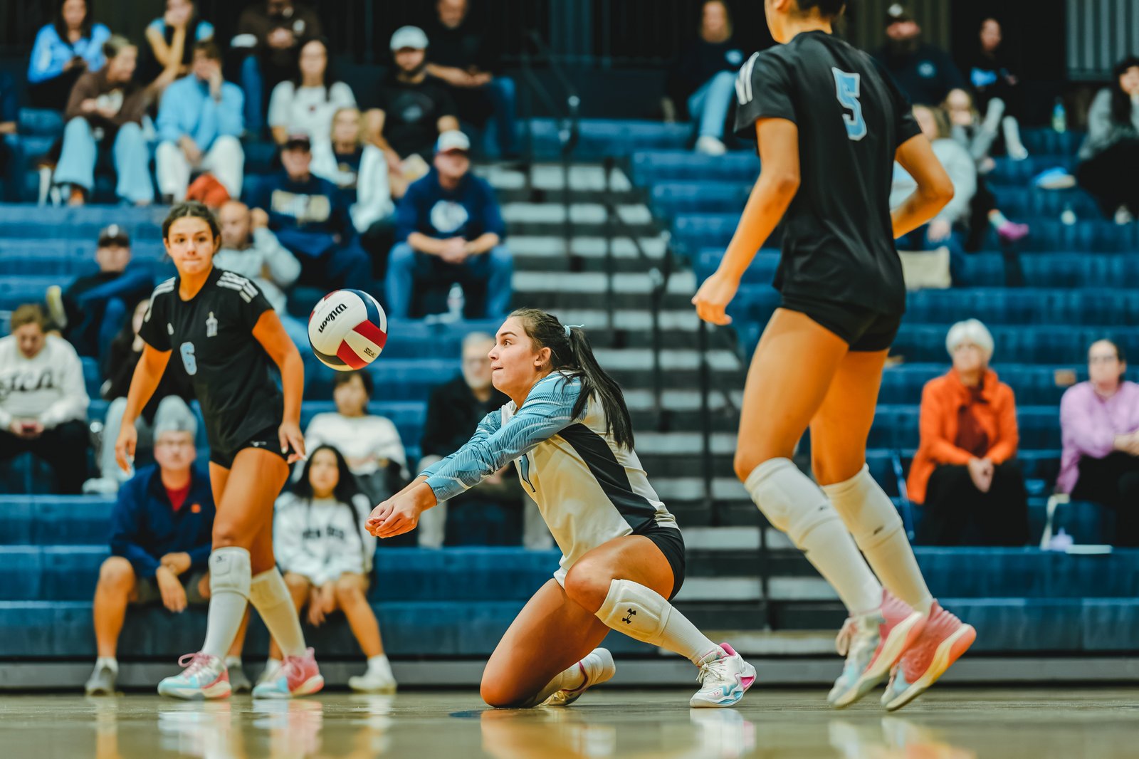 A player in a light blue top passes the ball; a player in black with number 5 and a player in black with number 6 are on court.