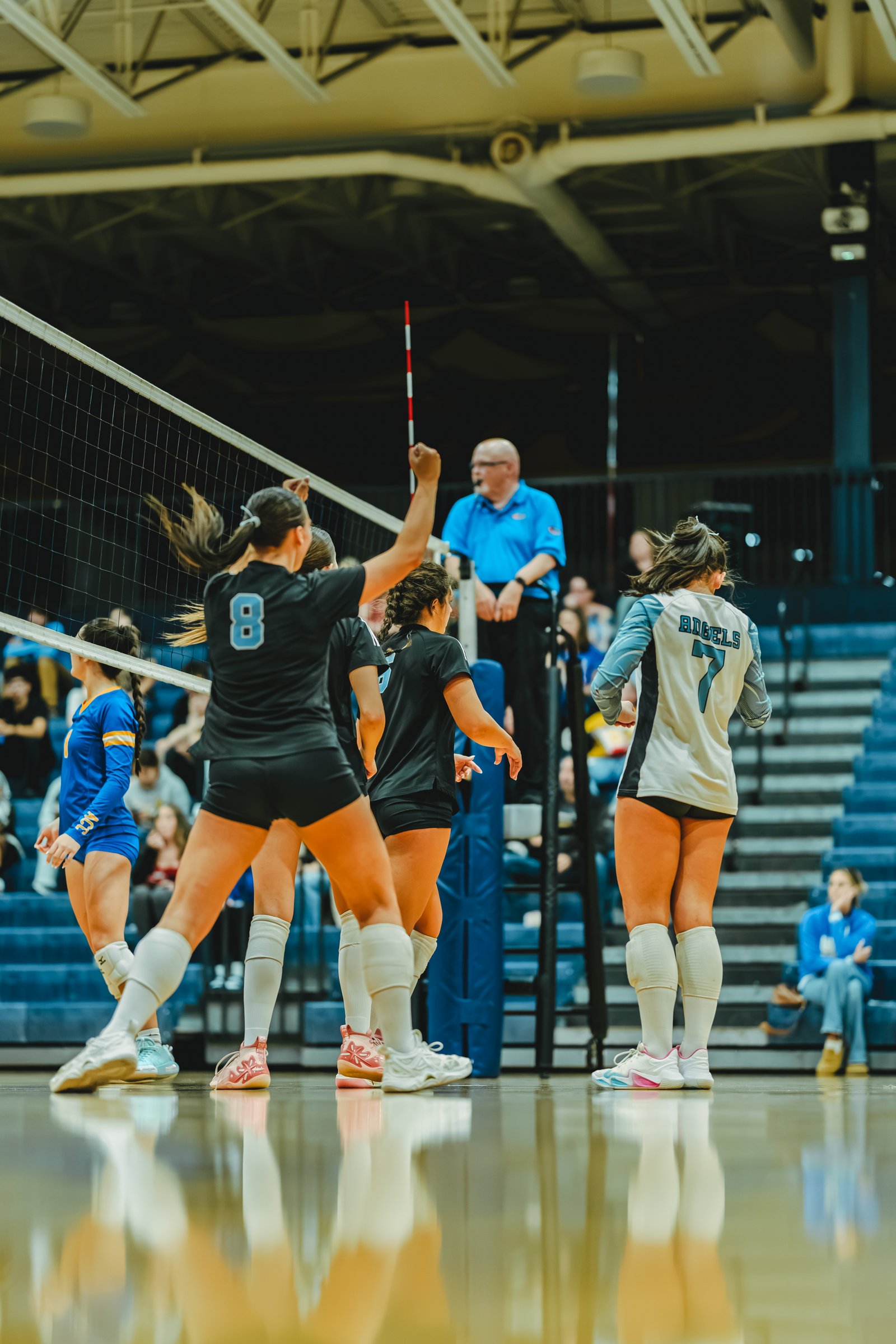 Players in black and light blue jerseys react near the net, with a referee watching. Player 8 in black and player 7 in light blue are visible.
