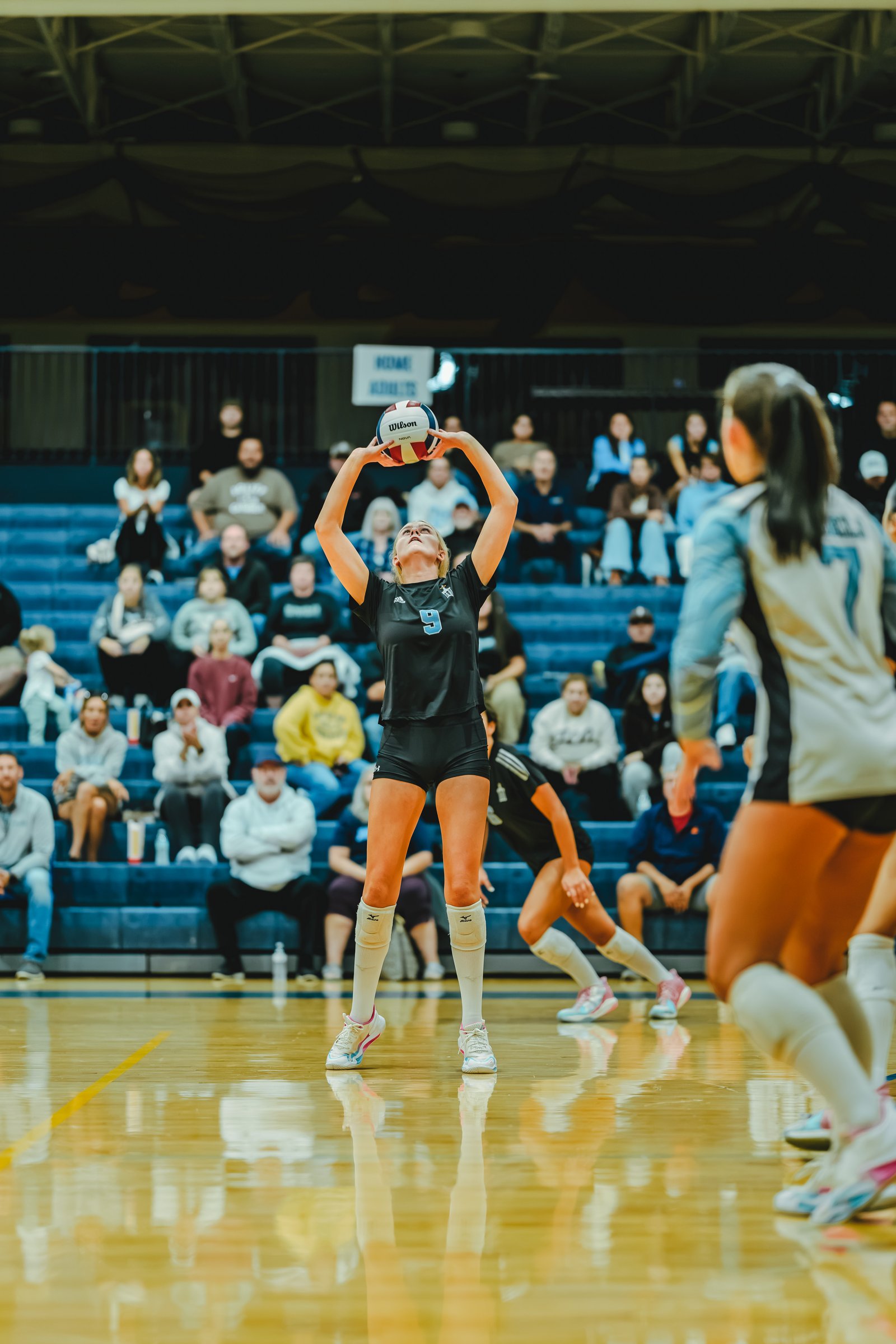 A player in a black top with number 9 prepares to serve a volleyball, with a player in a gray top visible on the right.