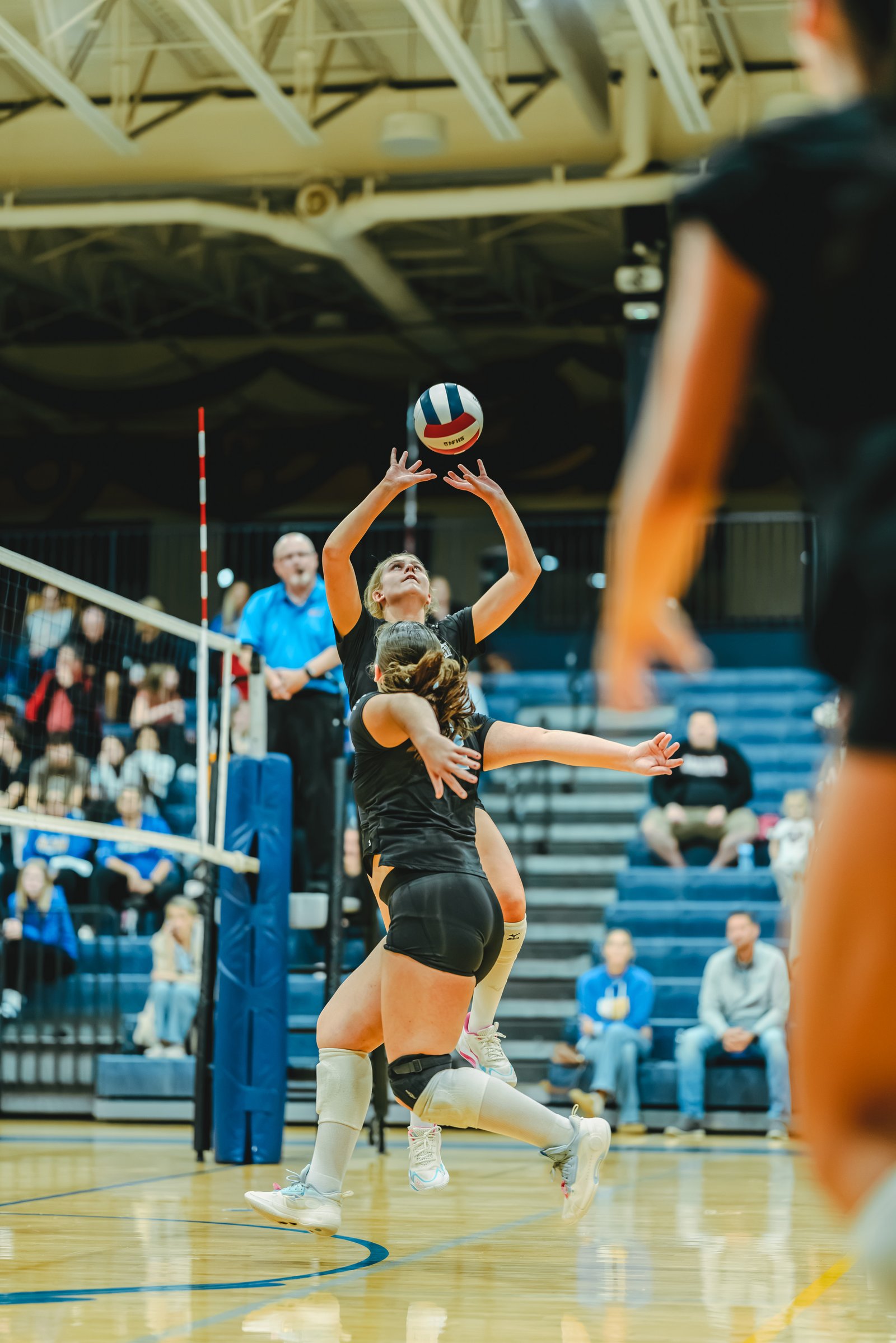 A player in black sets the ball while another player in black jumps to block during a volleyball game.