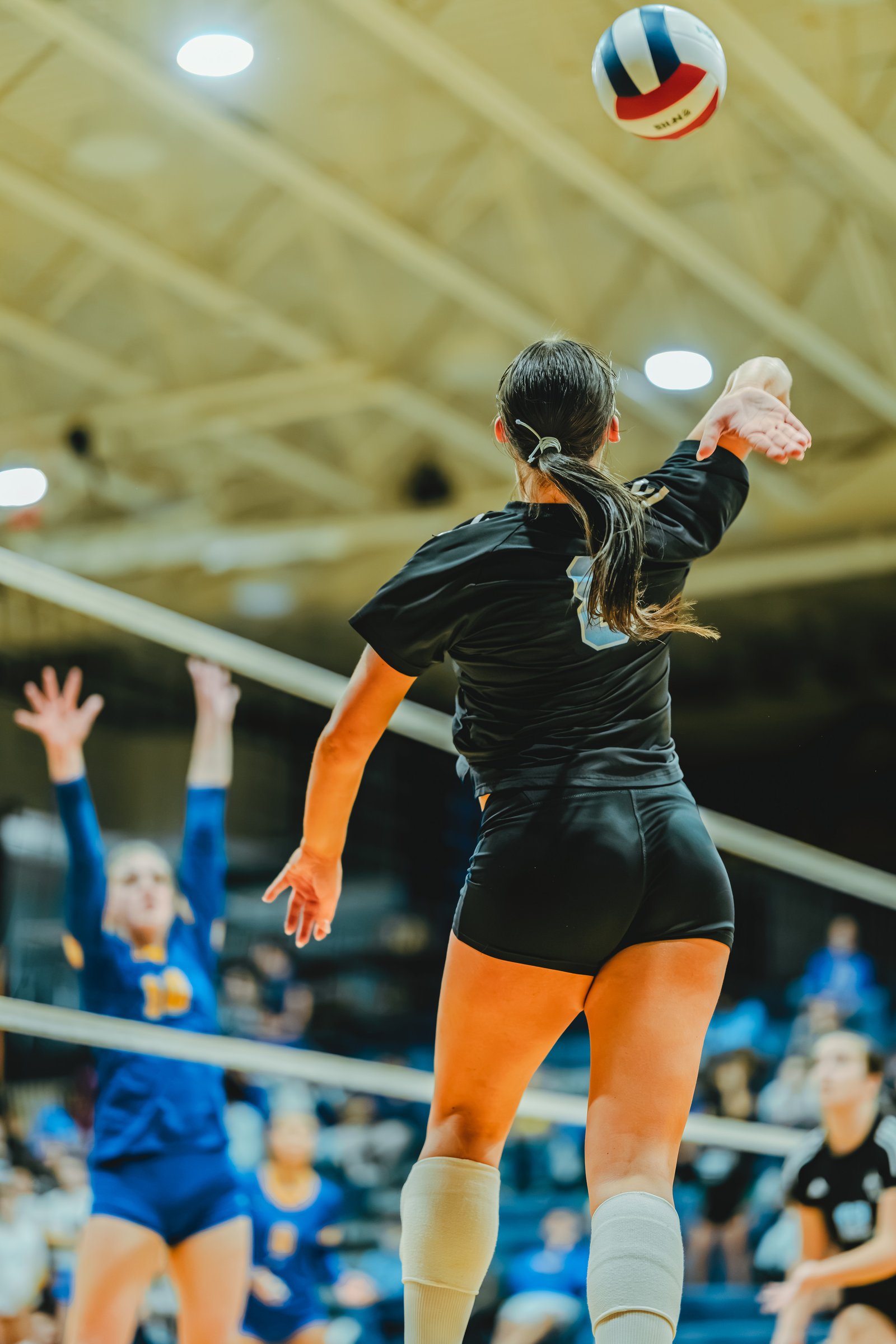 A player in black serves the volleyball as players in blue attempt to block. Player 10 in blue is visible.