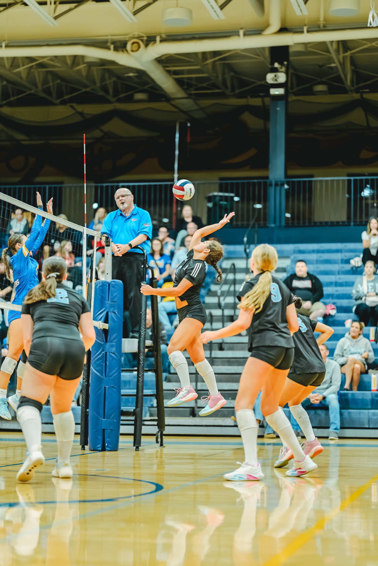 A player in black jumps to hit the ball, with players in black and blue on the court and a referee watching.