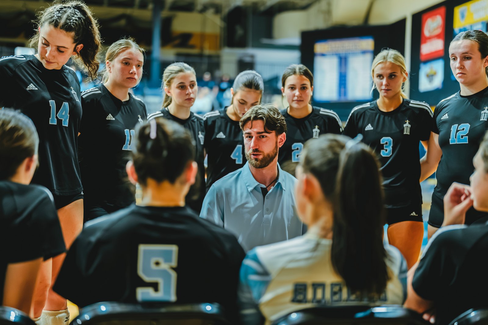 A coach talks to players in black jerseys, including number 14, number 11, number 4, number 3, and number 12, during a huddle.