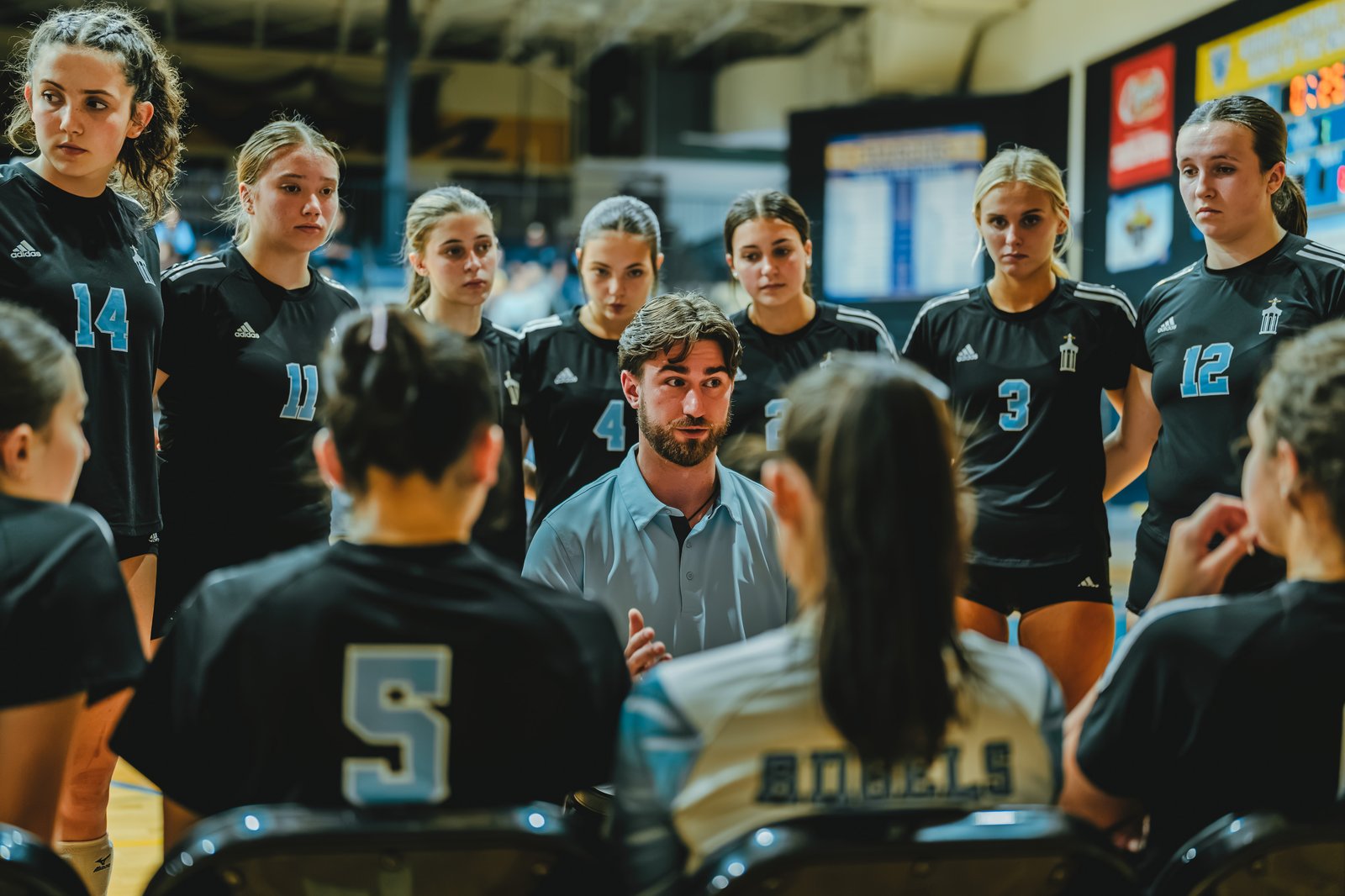 A coach talks to players in black jerseys, including number 14, 11, 4, 3, and 12, during a huddle.
