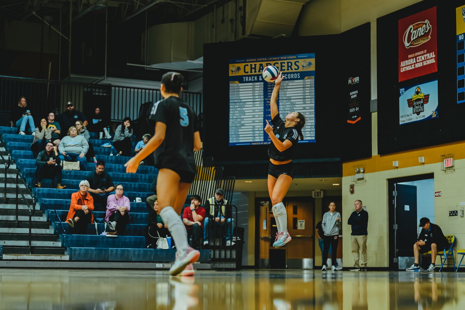 A player in black with white stripes jumps to hit a volleyball, while a player in black with a blue number 5 watches. Spectators are in the bleachers.
