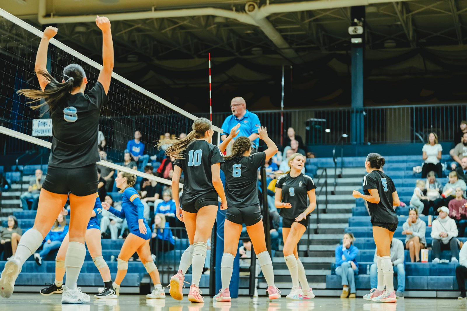 Players in black jerseys with numbers 8, 10, 6, 9, and 5, and a player in a blue jersey, are on the court during a volleyball game.