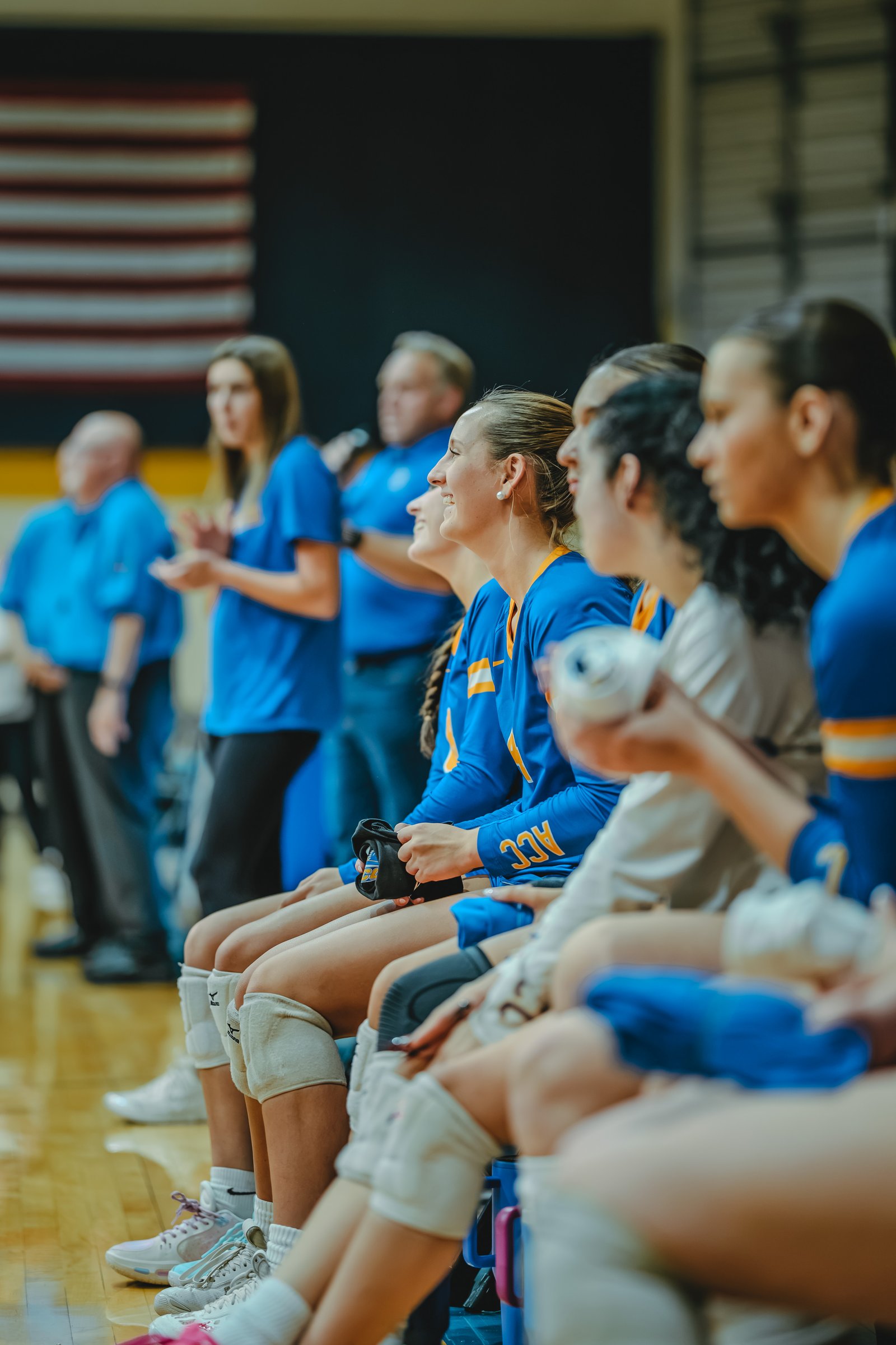 Players in blue and gold jerseys sit on the bench, with one player in a blue jersey holding a water bottle. ACC is visible on a jersey.