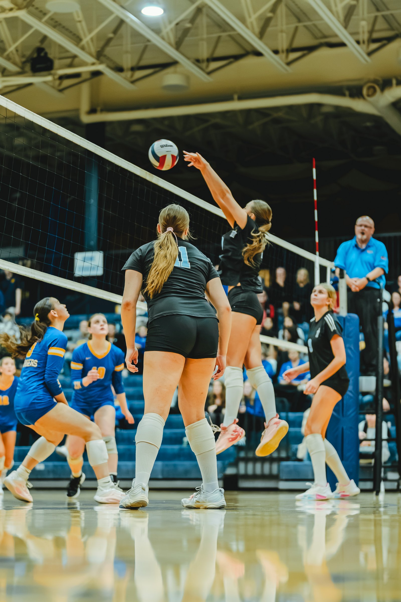 A player in black jumps to spike the ball, while players in blue and black watch. Player 11 in black and player 10 in blue are visible.