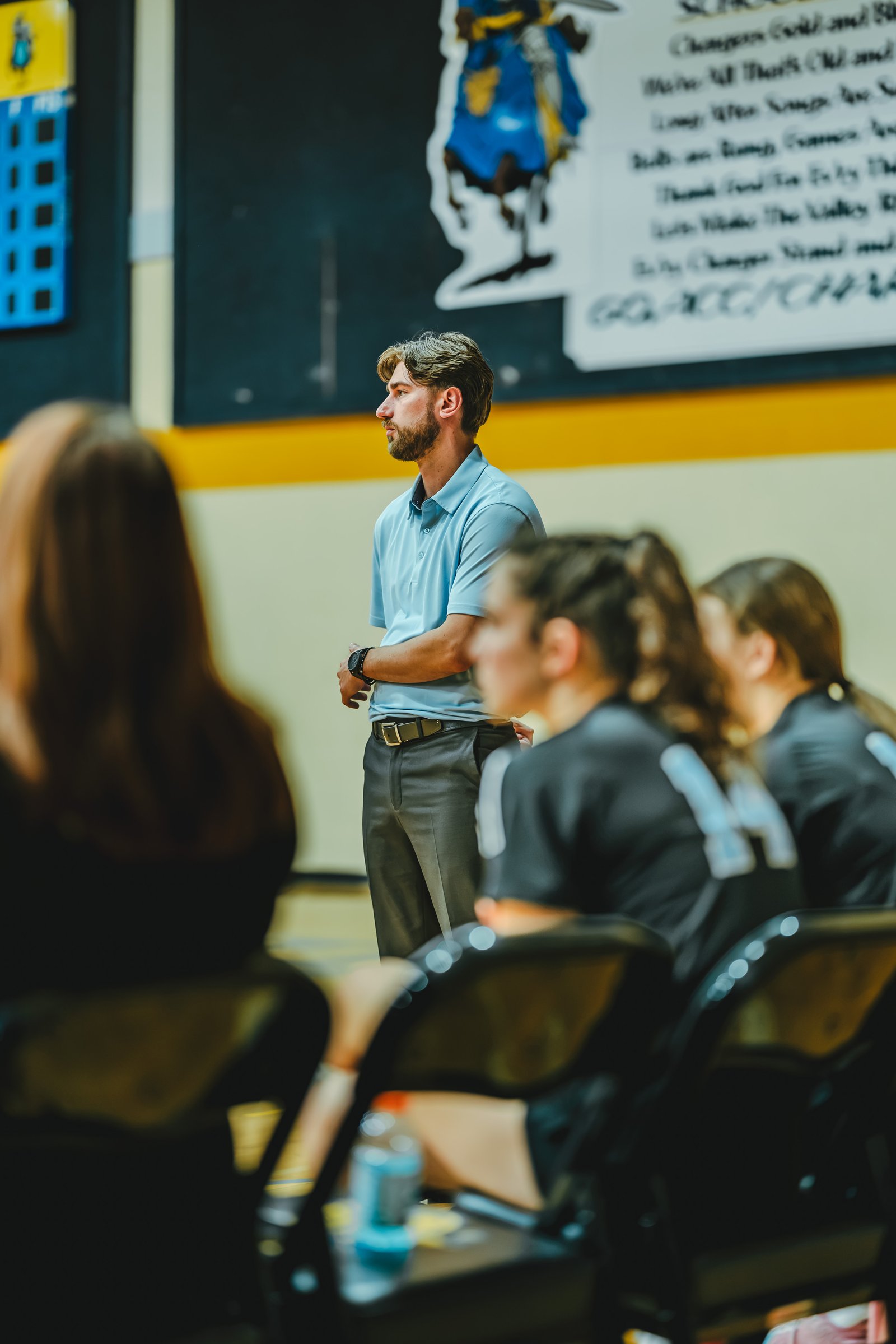 A coach watches players in black jerseys with white numbers, including player 14, during a volleyball game.