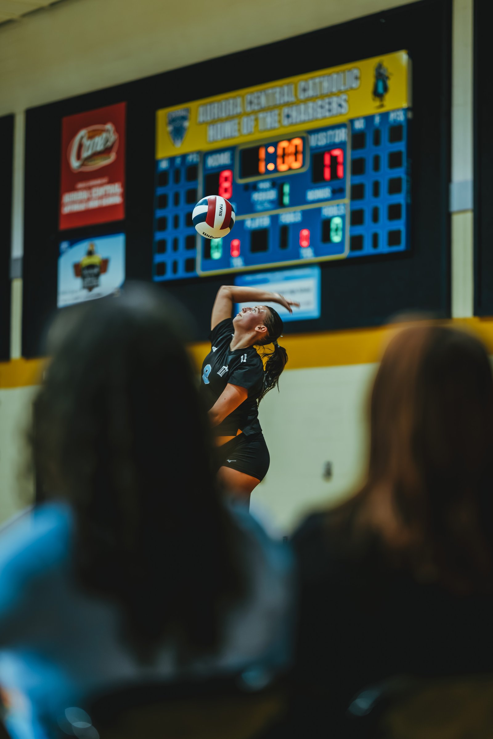 A player in a black top with number 11 serves the volleyball during a game, with a scoreboard in the background.