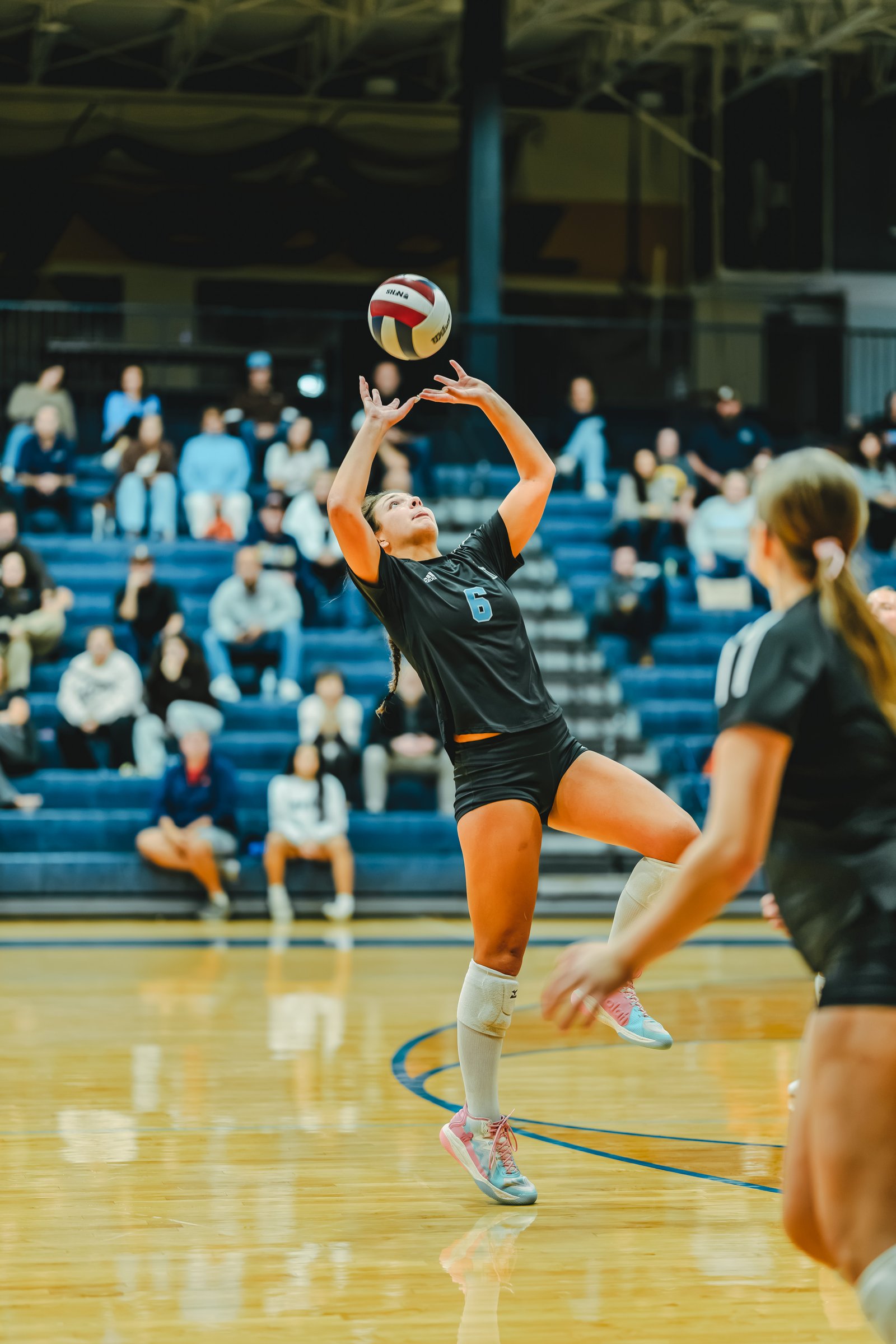 A player in a black top with number 6 sets the volleyball as another player in a black top with white stripes watches.