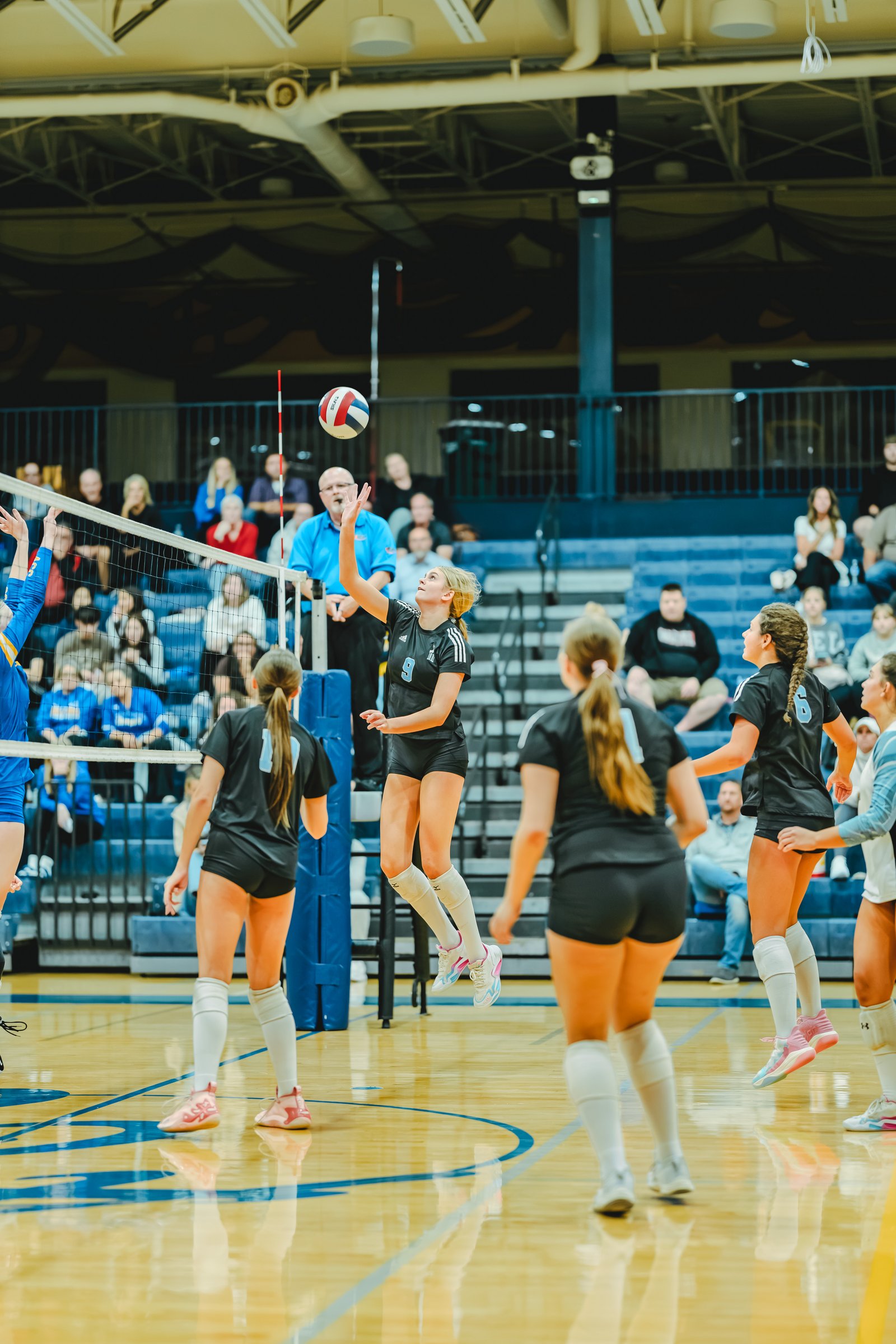 Player #9 in black jumps to hit the volleyball, with players #10 and #6 in black and a player in blue on the court.