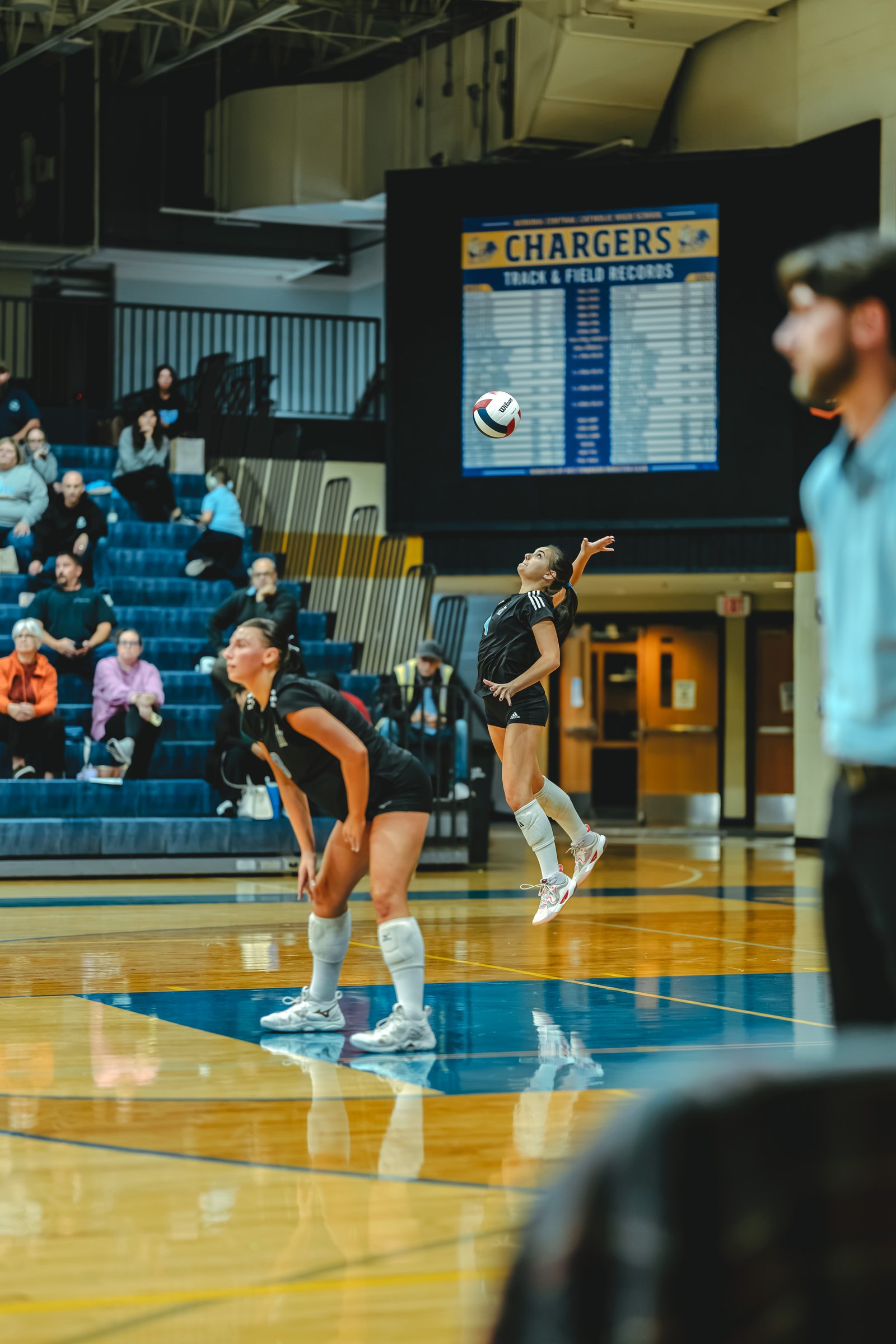 A player in black with number 4 jumps to serve a volleyball, while a player in black with number 12 prepares to receive.