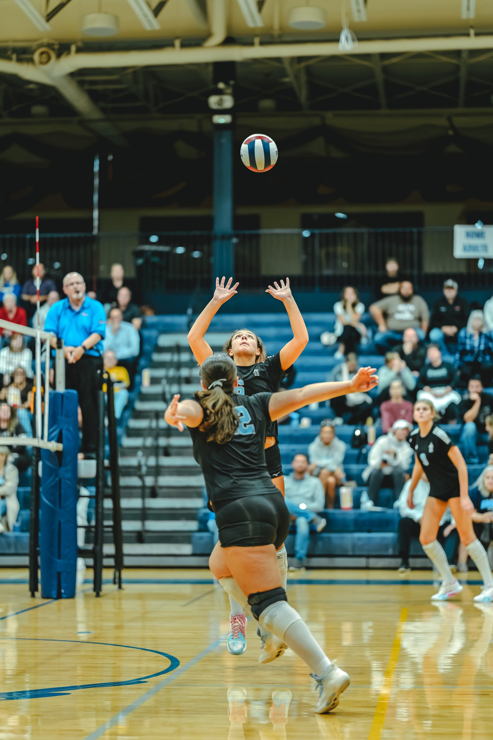 A player in black with jersey number 6 sets the ball as another player in black with jersey number 2 attempts to block, with spectators watching.