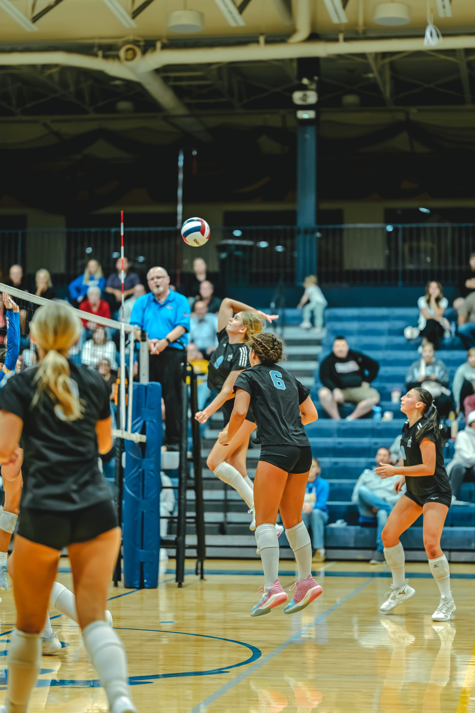 A player in black with jersey number 6 jumps to hit the ball, while a player in black with jersey number 3 watches. A player in blue serves.