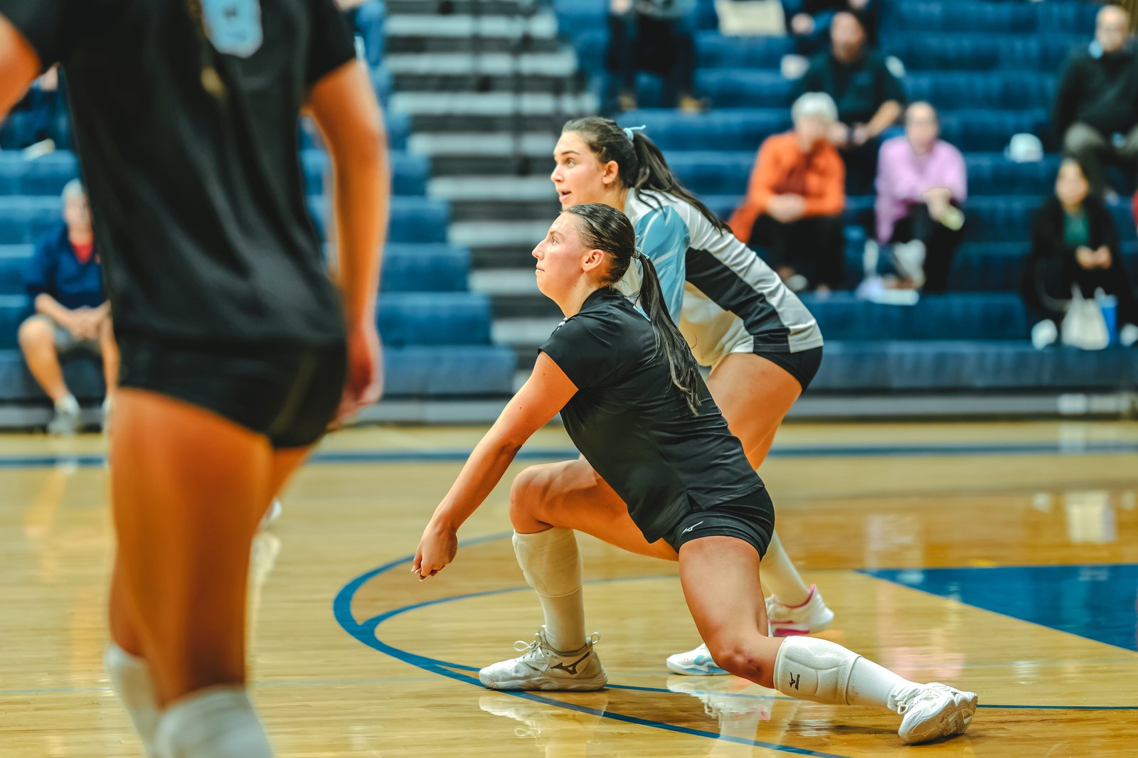 A player in black lunges for a volleyball, with a player in light blue and white behind her, and a player in black in the foreground.