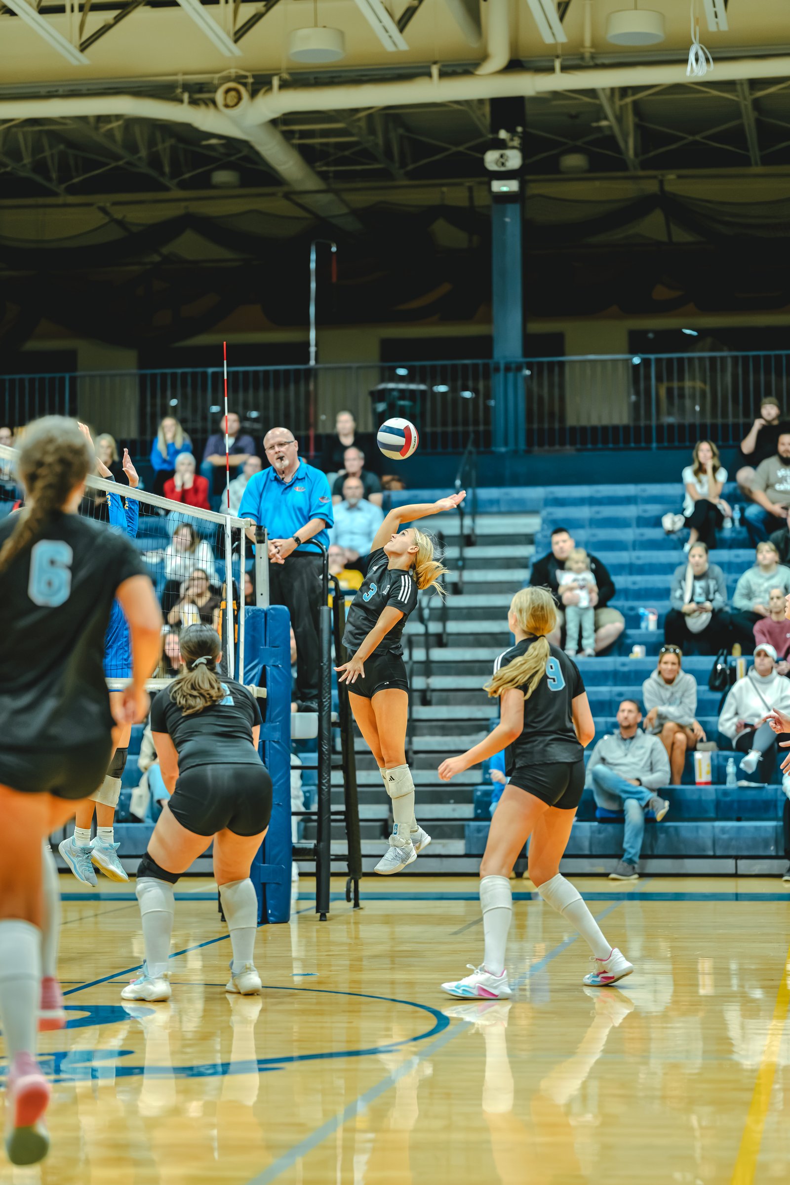 A player in black with number 3 jumps to hit the ball, while players in black with numbers 6 and 9 and a player in blue are on the court.