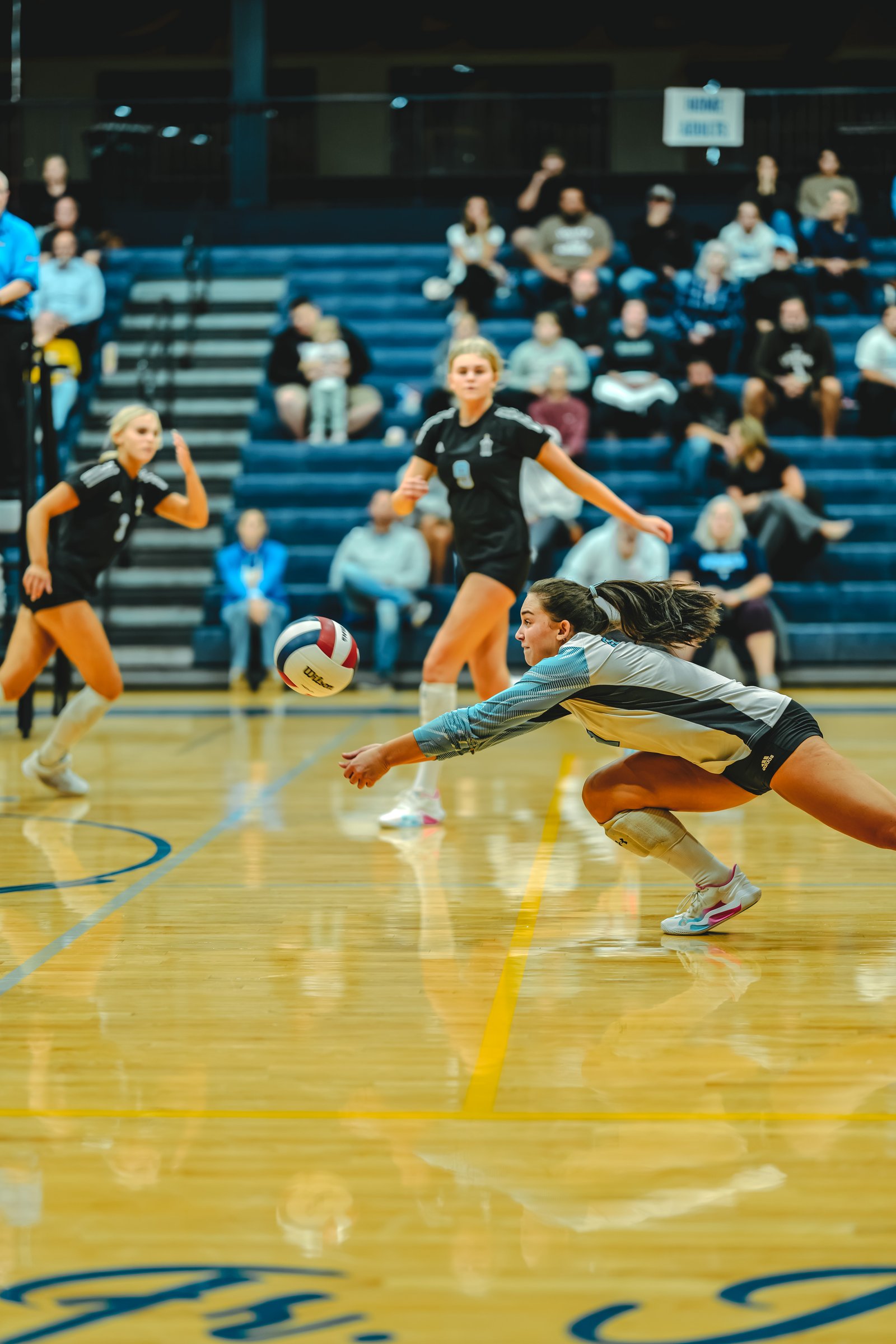 A player in a light blue and gray top dives for a volleyball, while a player in a black top with number 8 watches.