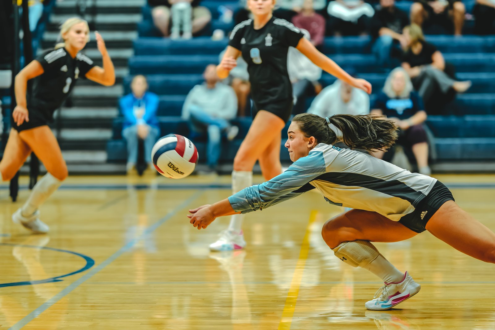 A player in a white and blue top dives for a volleyball, with players in black tops visible on the court.