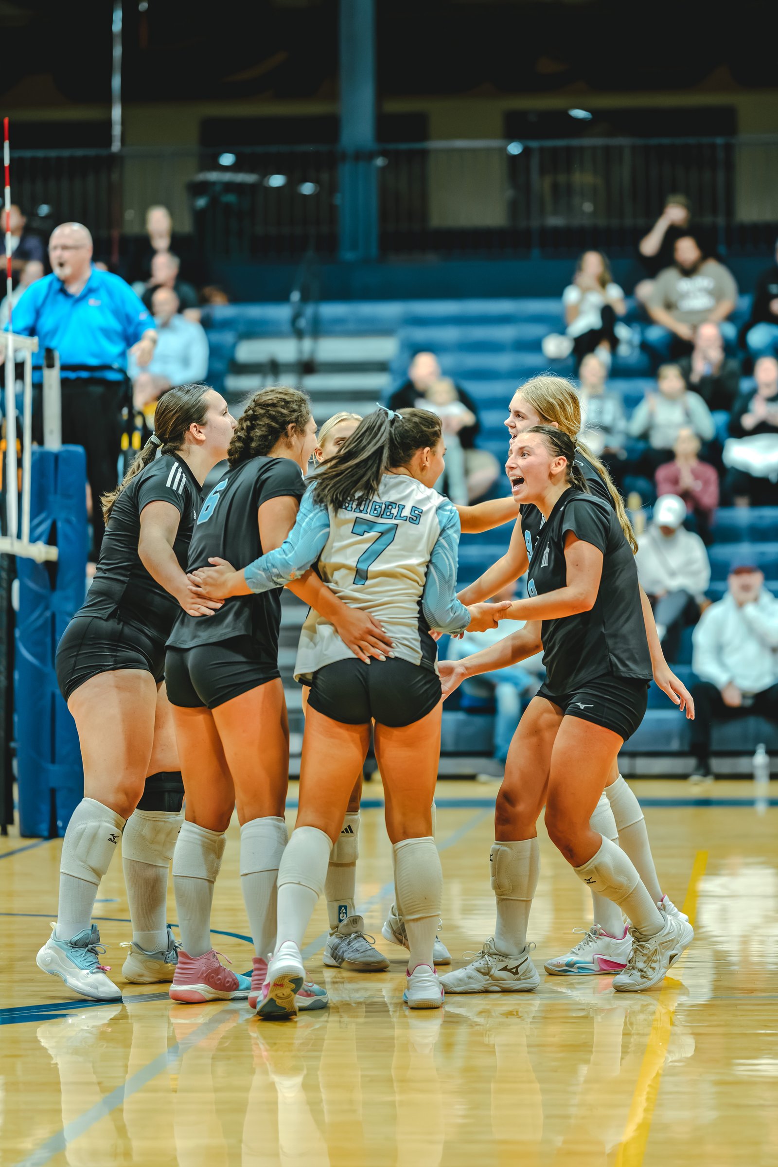 Players in black and light blue jerseys huddle together on the court, with player number 7 in the center. Player number 9 is visible in black.