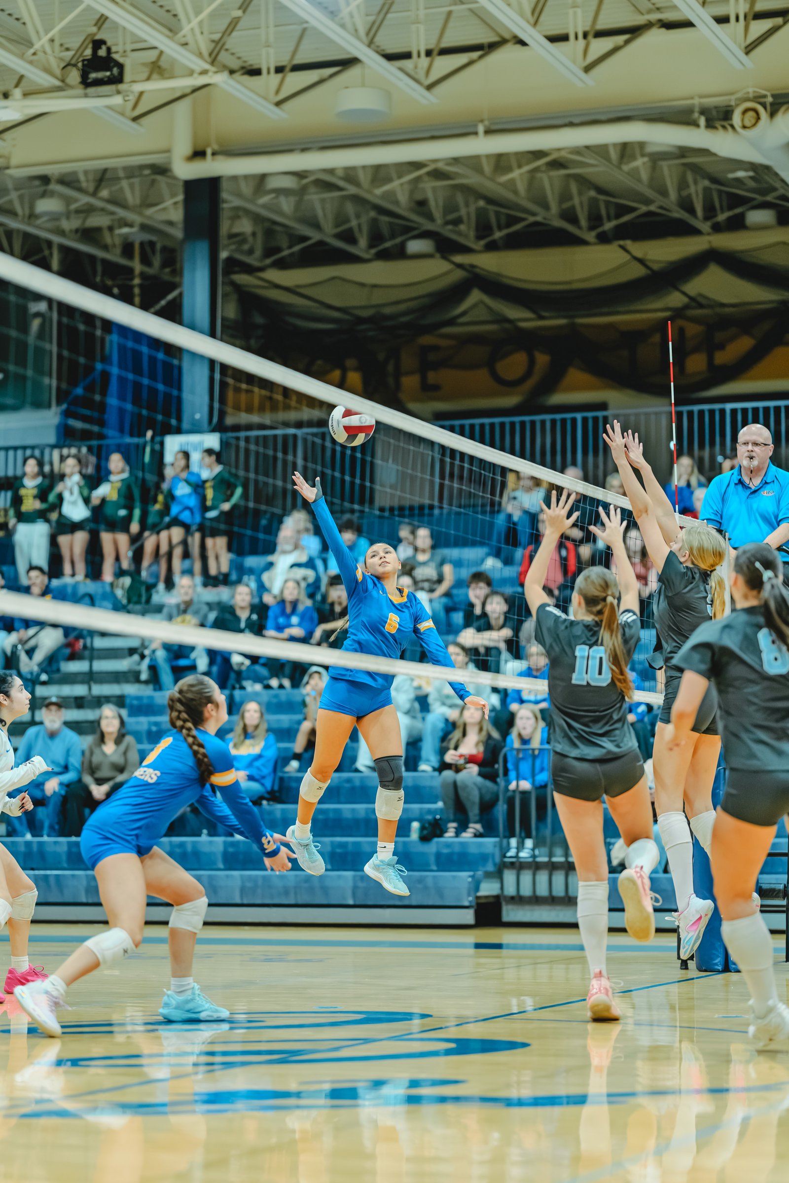 A player in blue with jersey number 6 jumps to hit the ball, while players in black with numbers 10 and 8 attempt to block.