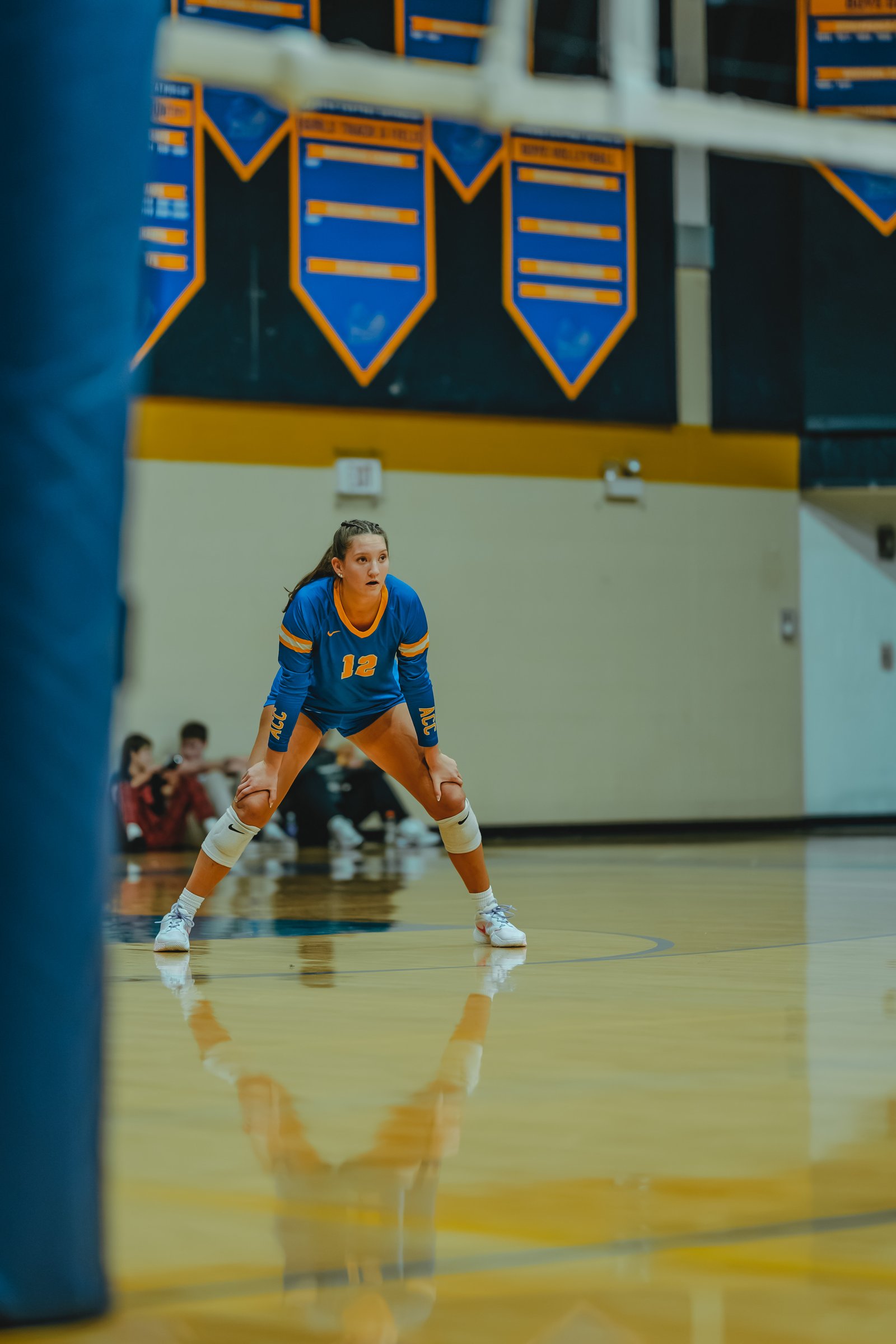 A player in a blue jersey with orange trim and number 12 crouches on the court, with other players blurred in the background.