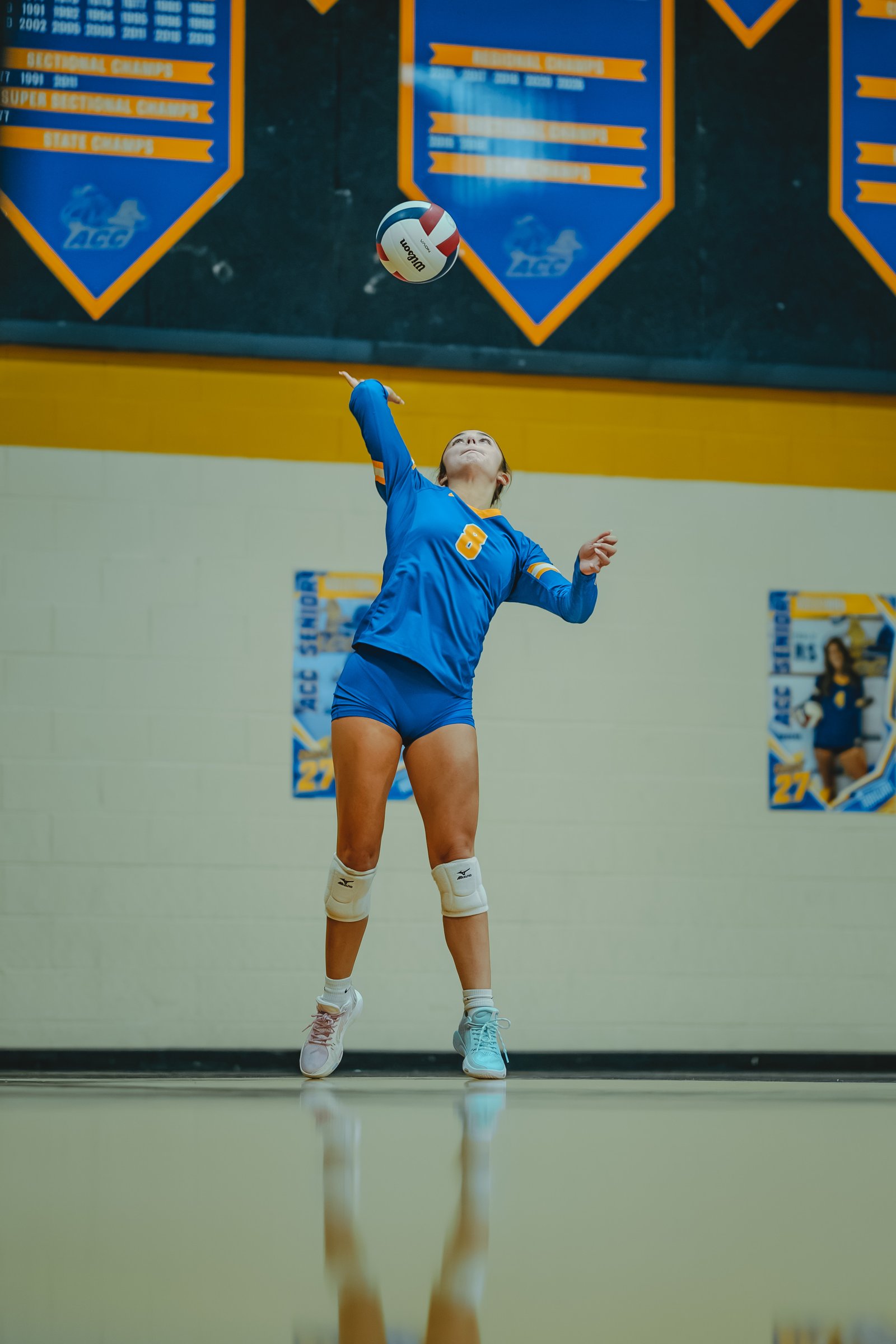 A player in a blue jersey with orange trim, number 8, serves the volleyball. Another player in blue is visible in the background.