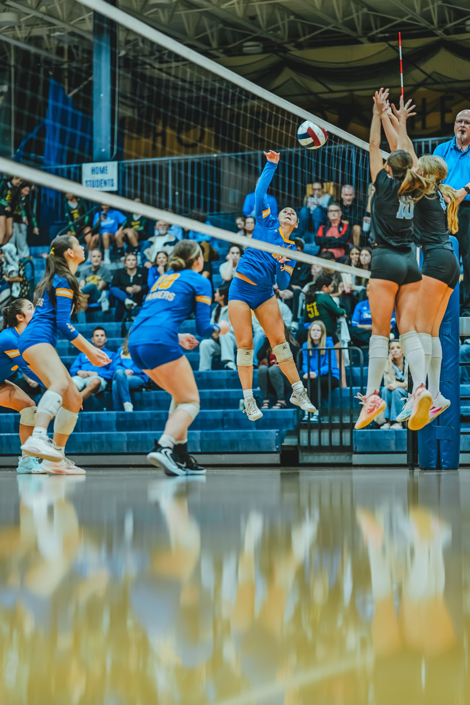 A player in blue spikes the ball as players in black and blue jump to block, with a player in blue in the foreground.