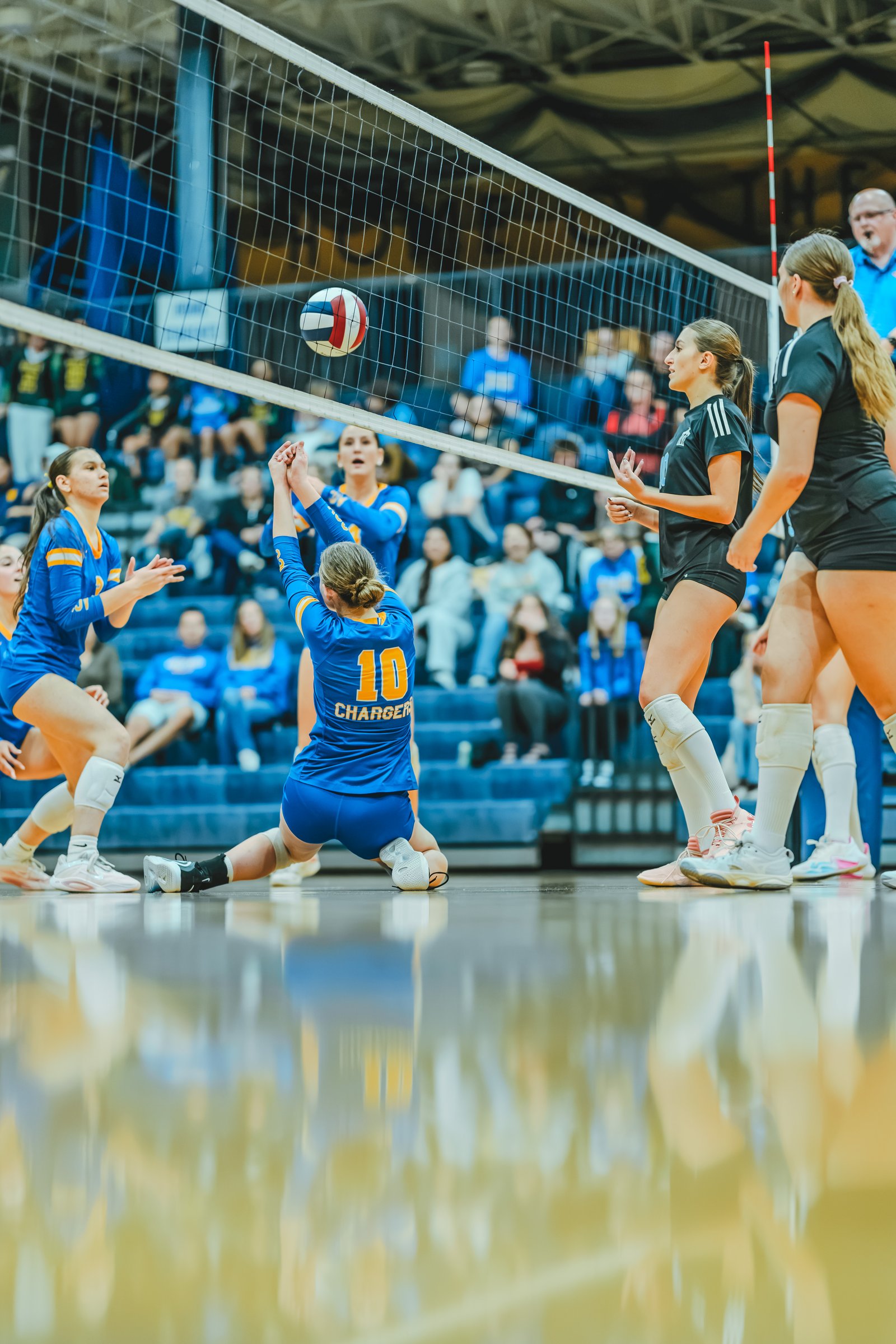A player in blue with jersey number 10 dives for the ball as a player in black watches, with other players in blue and black on the court.