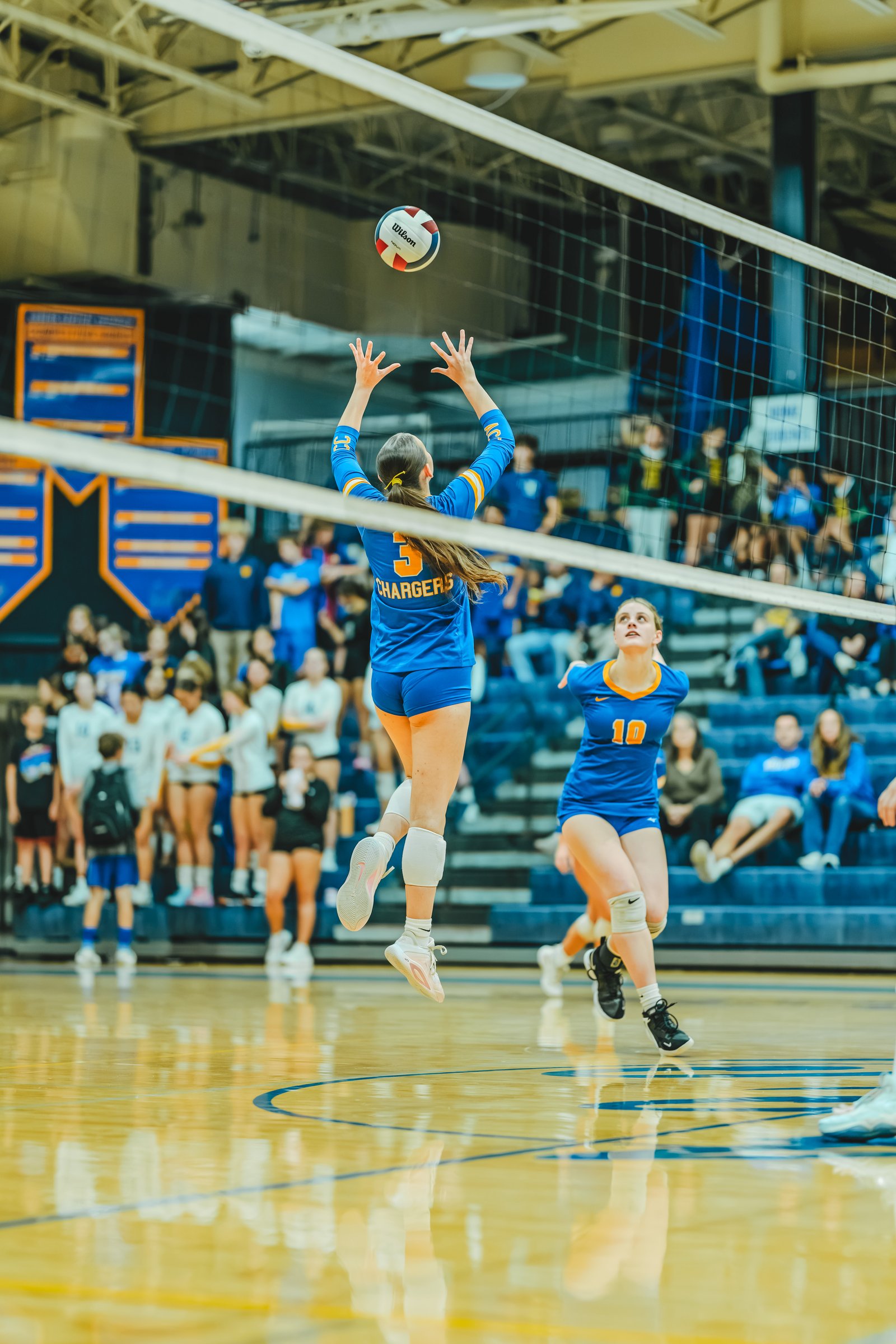 A player in blue with jersey number 5 jumps to hit the ball, while a player in blue with jersey number 10 watches.