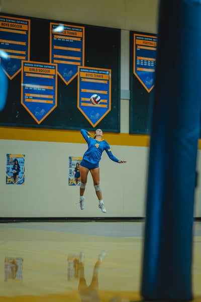 A player in a blue jersey with number 4 jumps to serve the volleyball. Another player in a blue jersey with number 27 is visible on a poster.
