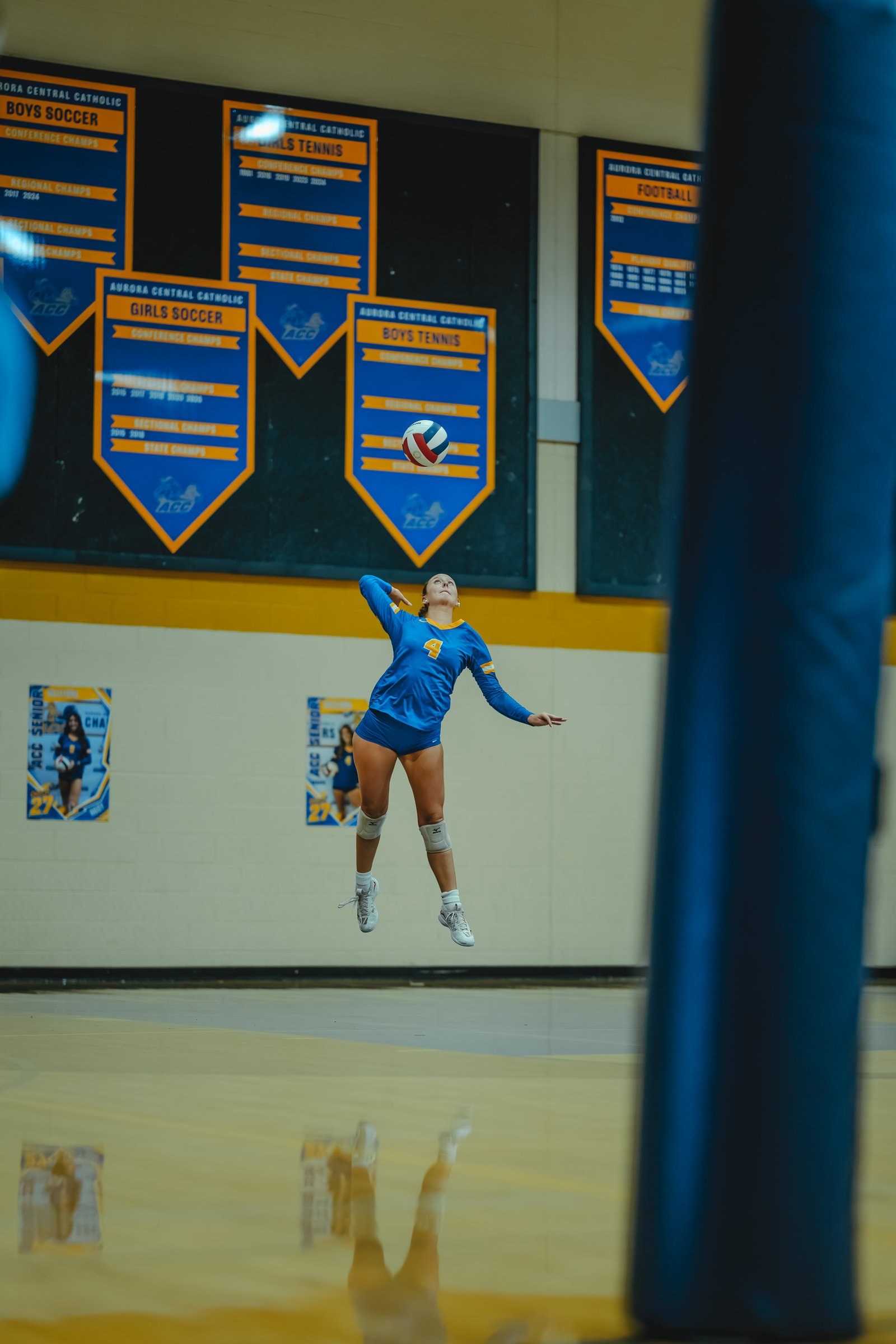 A player in a blue jersey with number 4 jumps to serve the volleyball. Another player in a blue jersey with number 27 is visible on a poster.