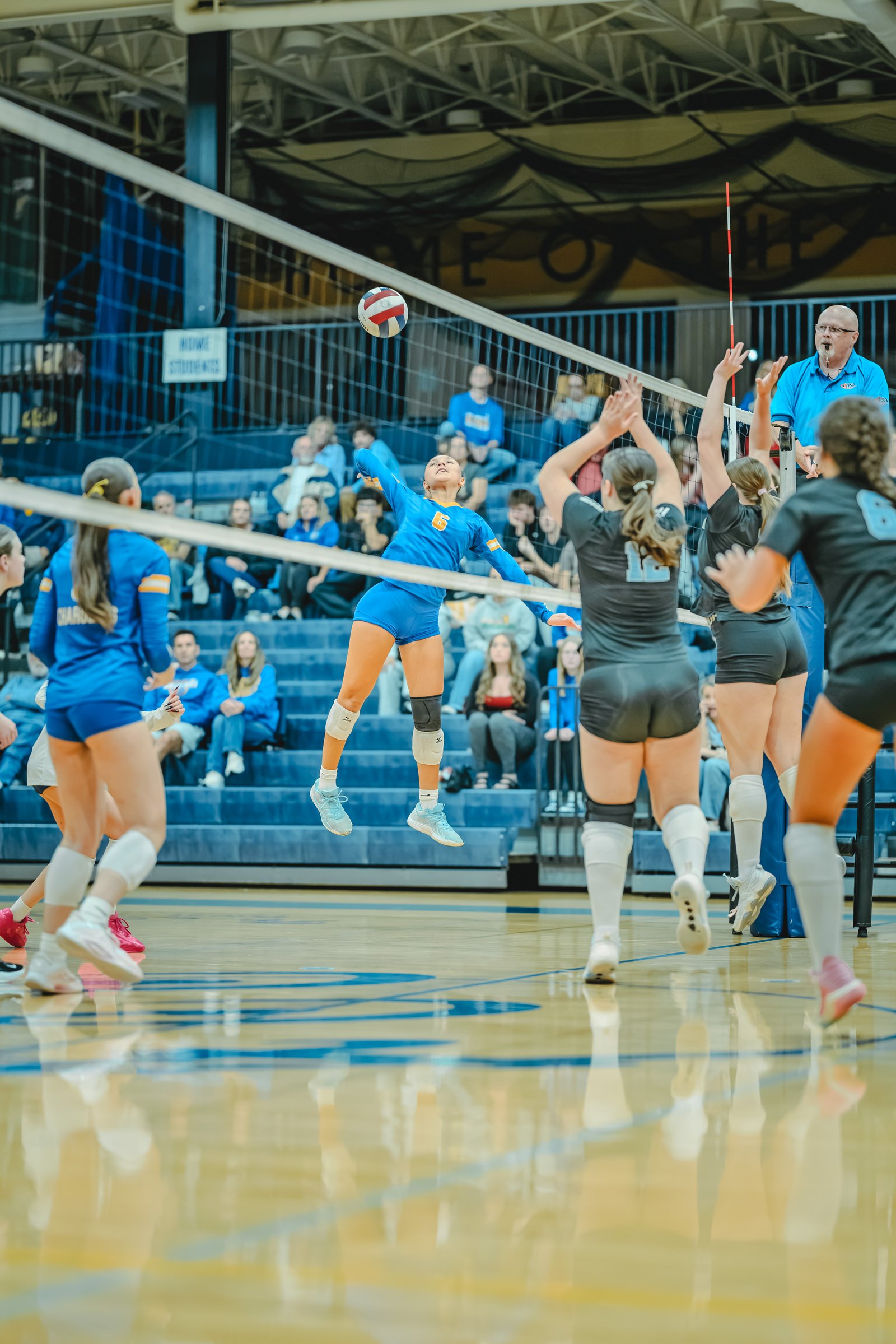 A player in blue with jersey number 6 jumps to hit the volleyball as players in black with jersey numbers 12 and 15 attempt to block.