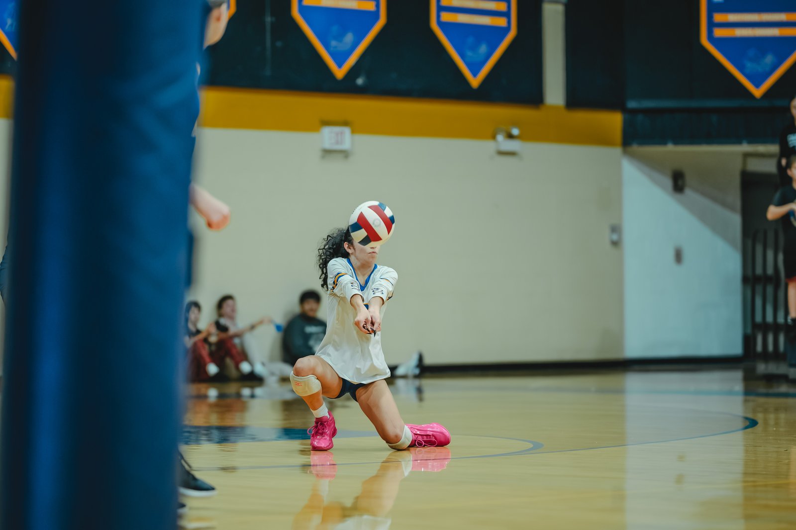 A player in white with blue and gold trim dives to pass the ball. Another player in black sits on the bench.