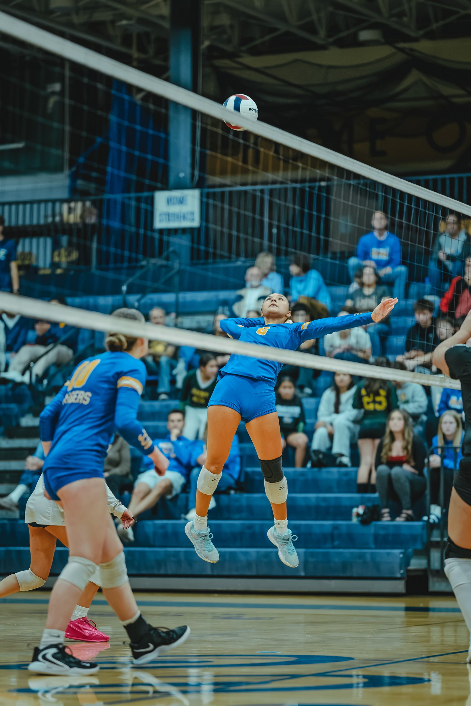 A player in blue jumps to hit the volleyball, with a player in blue number 10 and a player in black visible on court.