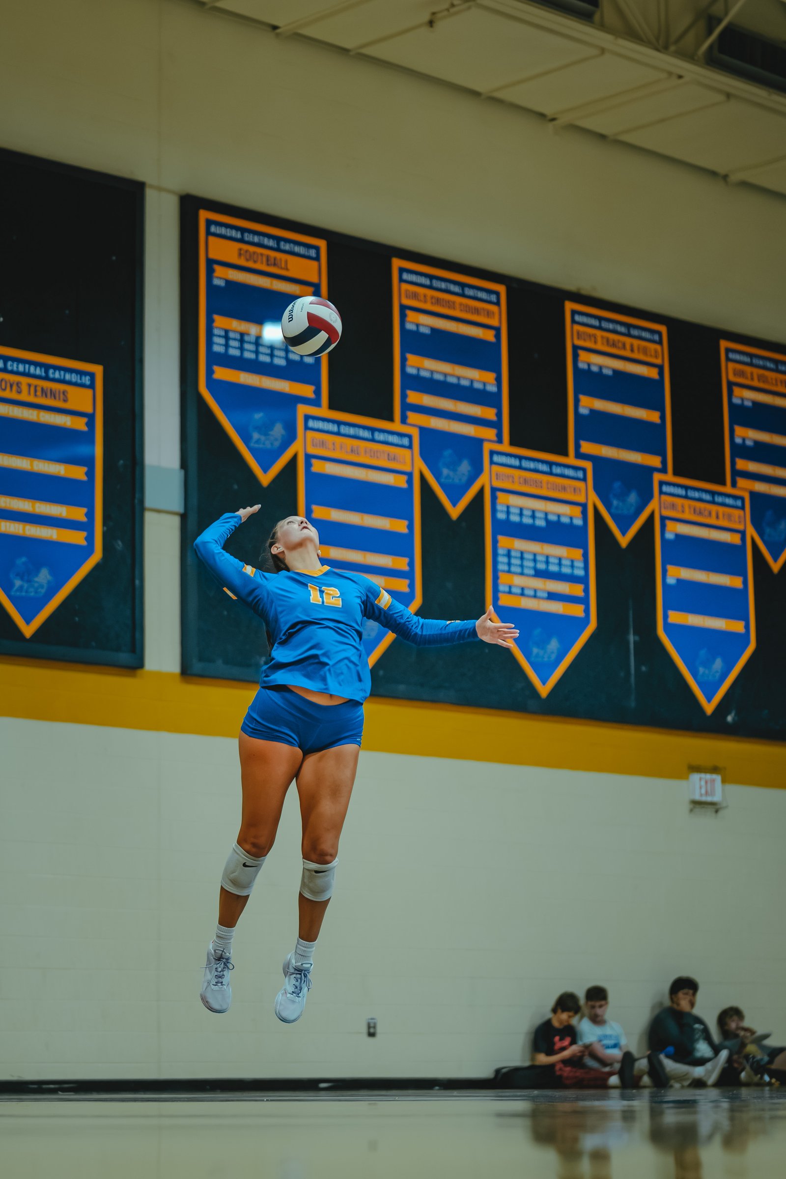 A player in a blue jersey with yellow trim, number 12, jumps to hit a volleyball. Other players sit on the floor in the background.