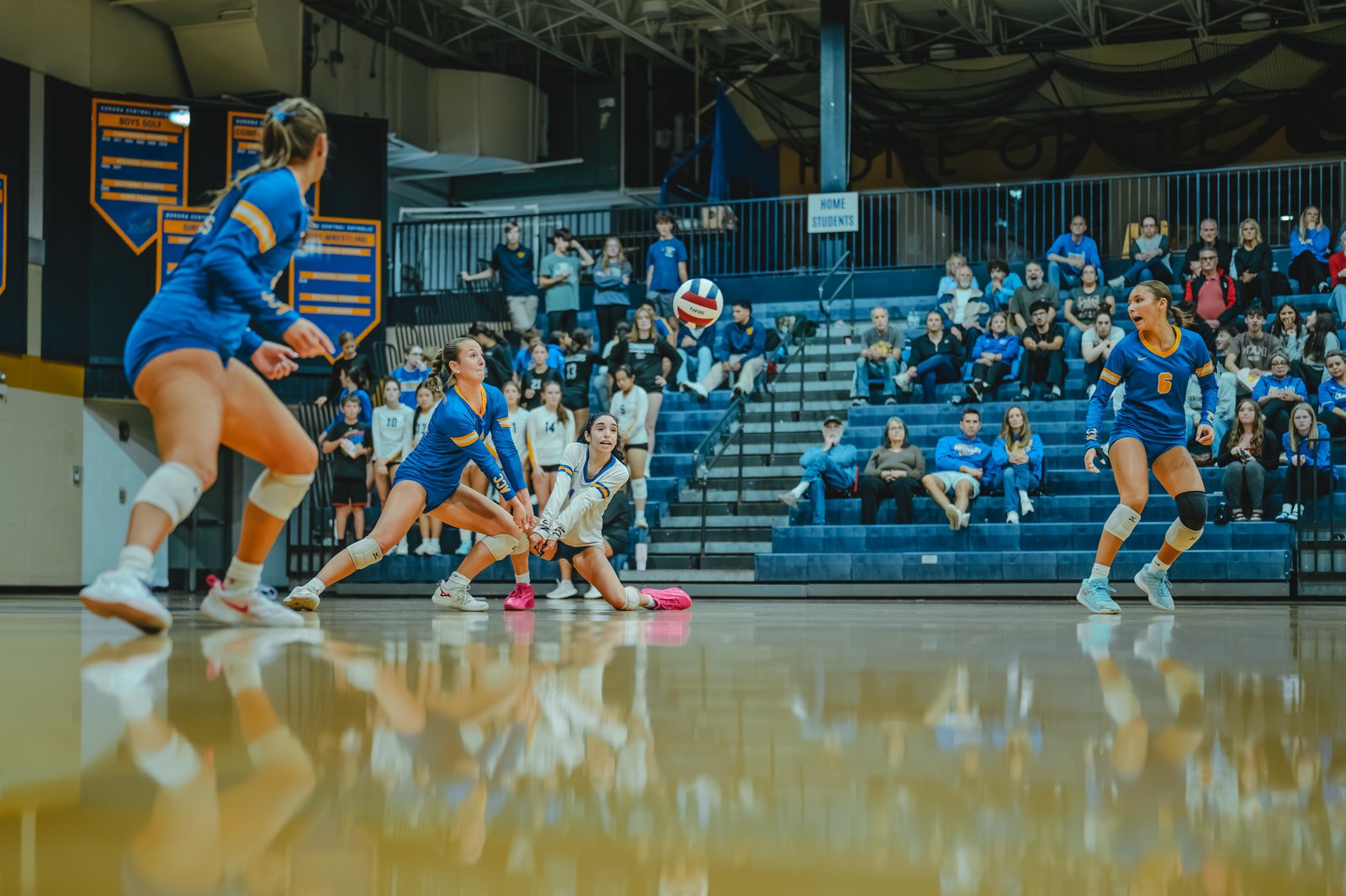 A player in blue with jersey number 6 and a player in white with jersey number 14 dive for the ball during a volleyball game.