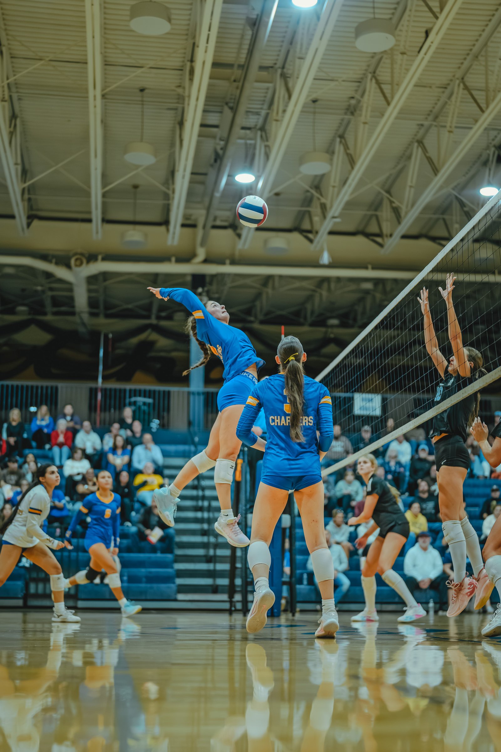 A player in blue jumps to spike the ball, with a player in blue number 5 and a player in black blocking at the net.