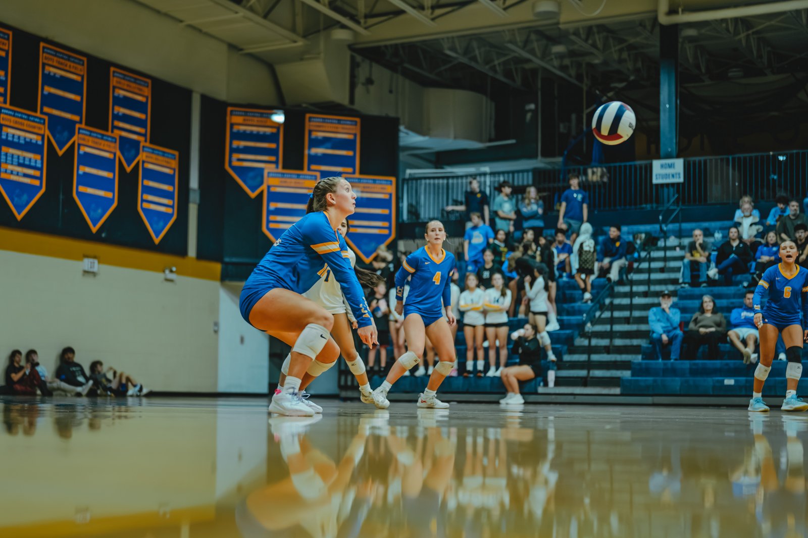 A player in blue with jersey number 4 prepares to receive a volleyball, while players in blue with numbers 1 and 6 are visible on the court.