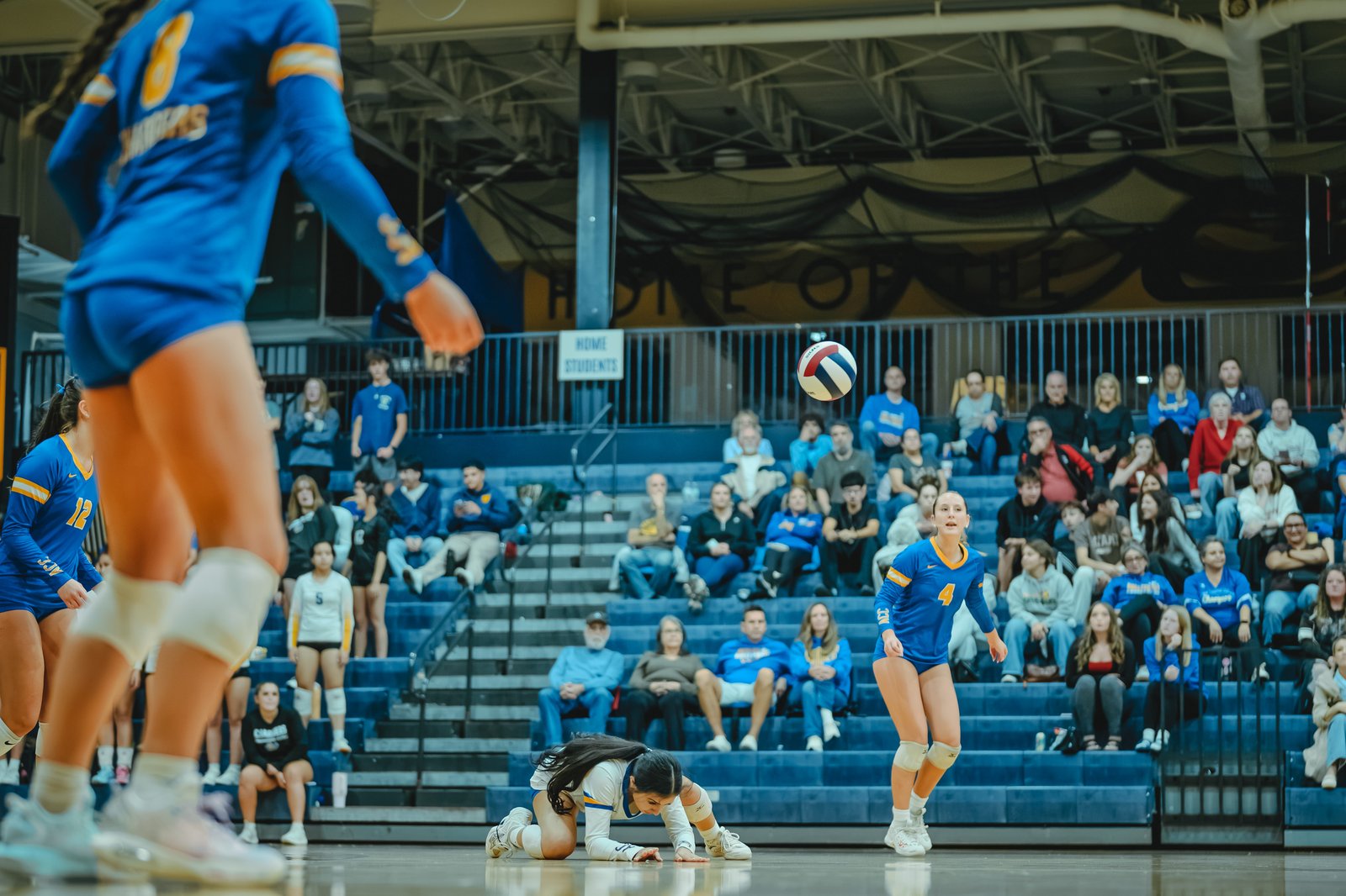 A player in blue with number 4 prepares to receive a volleyball, while players in blue number 12 and white number 5 are on court.