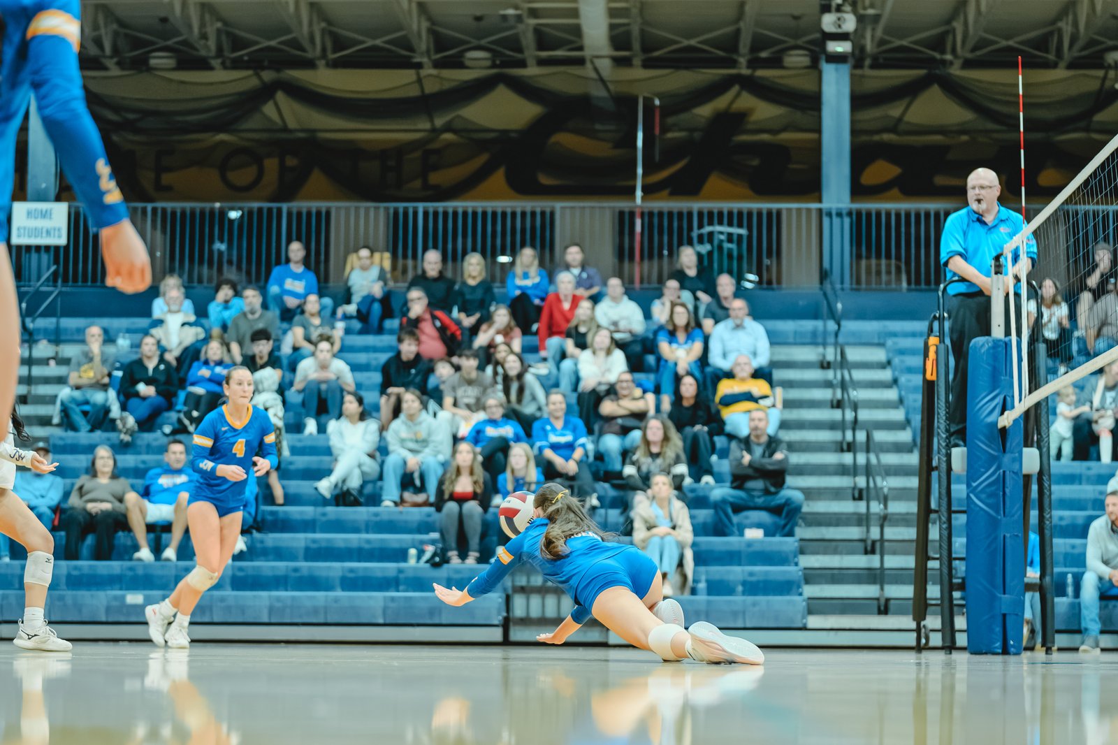 A player in blue dives for a volleyball, with a player in blue number 4 running nearby. Spectators watch from the bleachers.