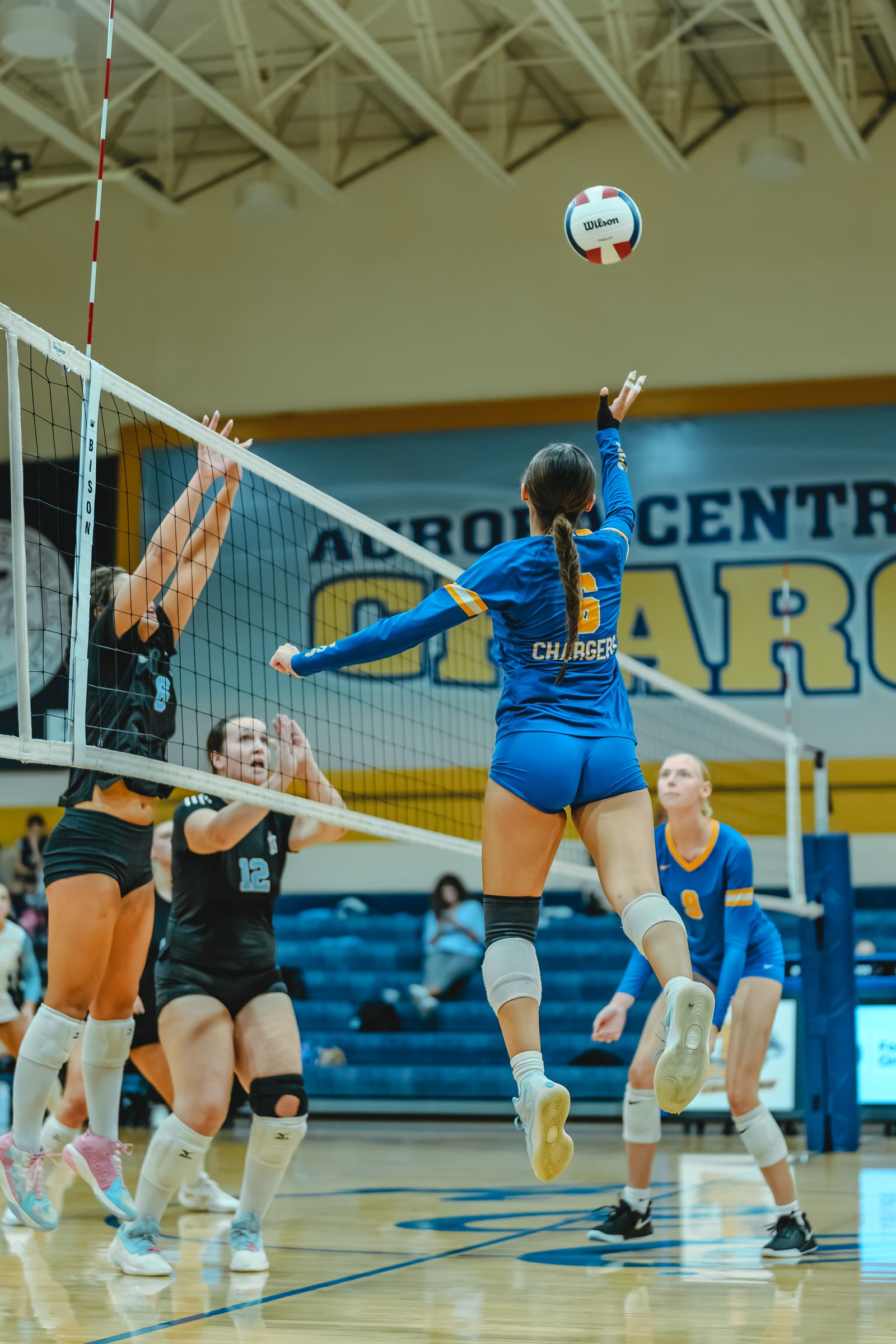 A player in blue with jersey number 5 spikes the ball over the net while players in black and blue defend.