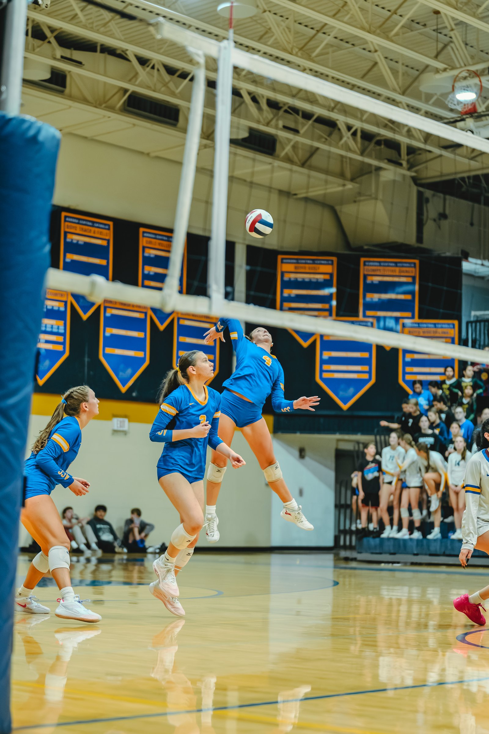 A player in blue jumps to spike the ball as teammates in blue and players in white watch during a volleyball game.